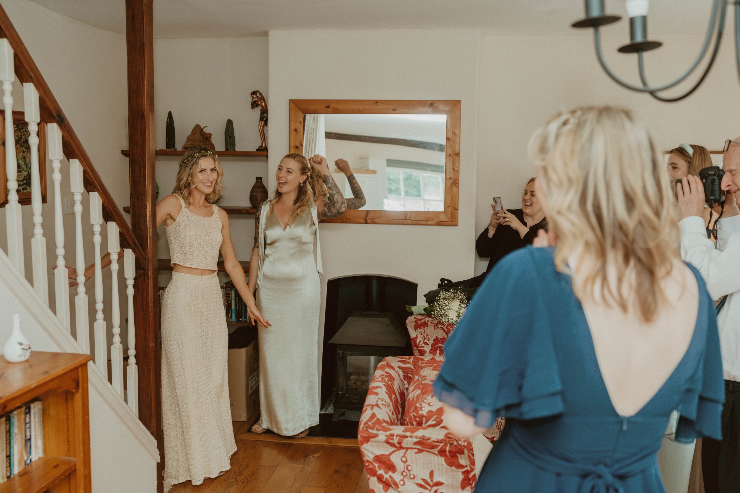 Two women, one in a cream-colored dress and the other in a satin gown, standing hand-in-hand in a living room, smiling, with friends taking photos and cheering.