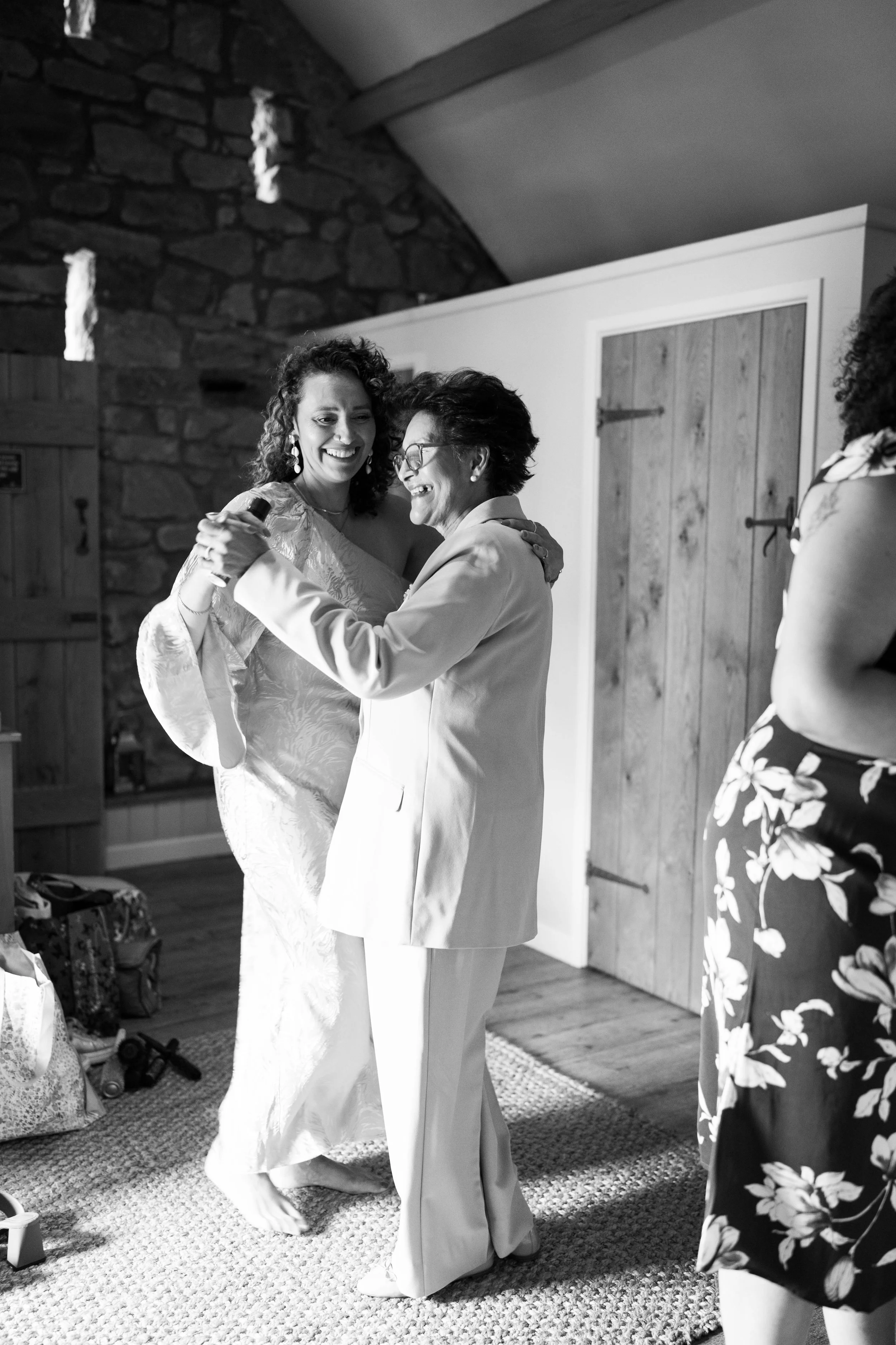 Two women dancing and smiling indoors, one in a wedding dress and the other in a light suit, with a third woman partially visible on the right side.