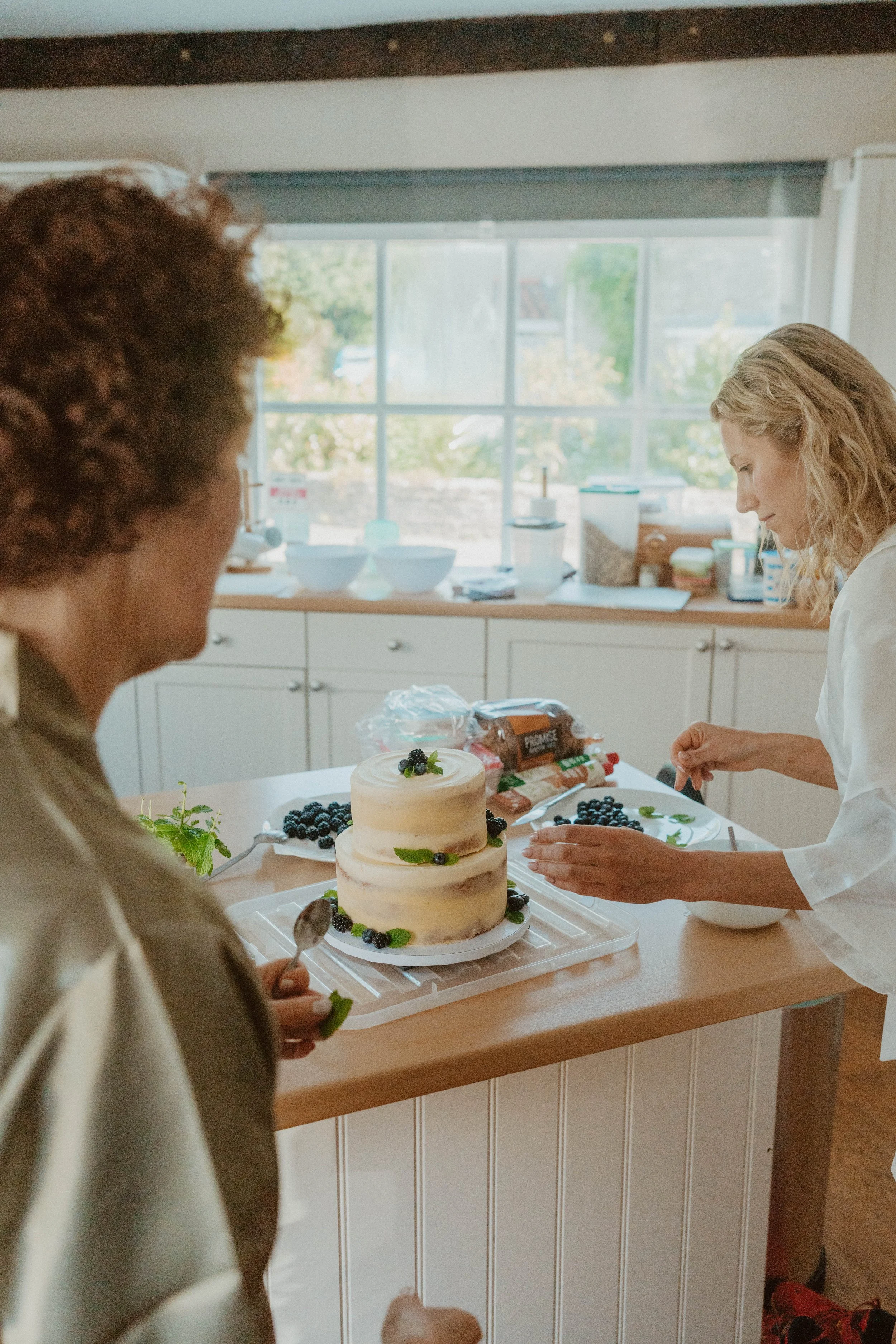 Two women decorating a two-tiered naked cake with berries and mint leaves in a bright kitchen.