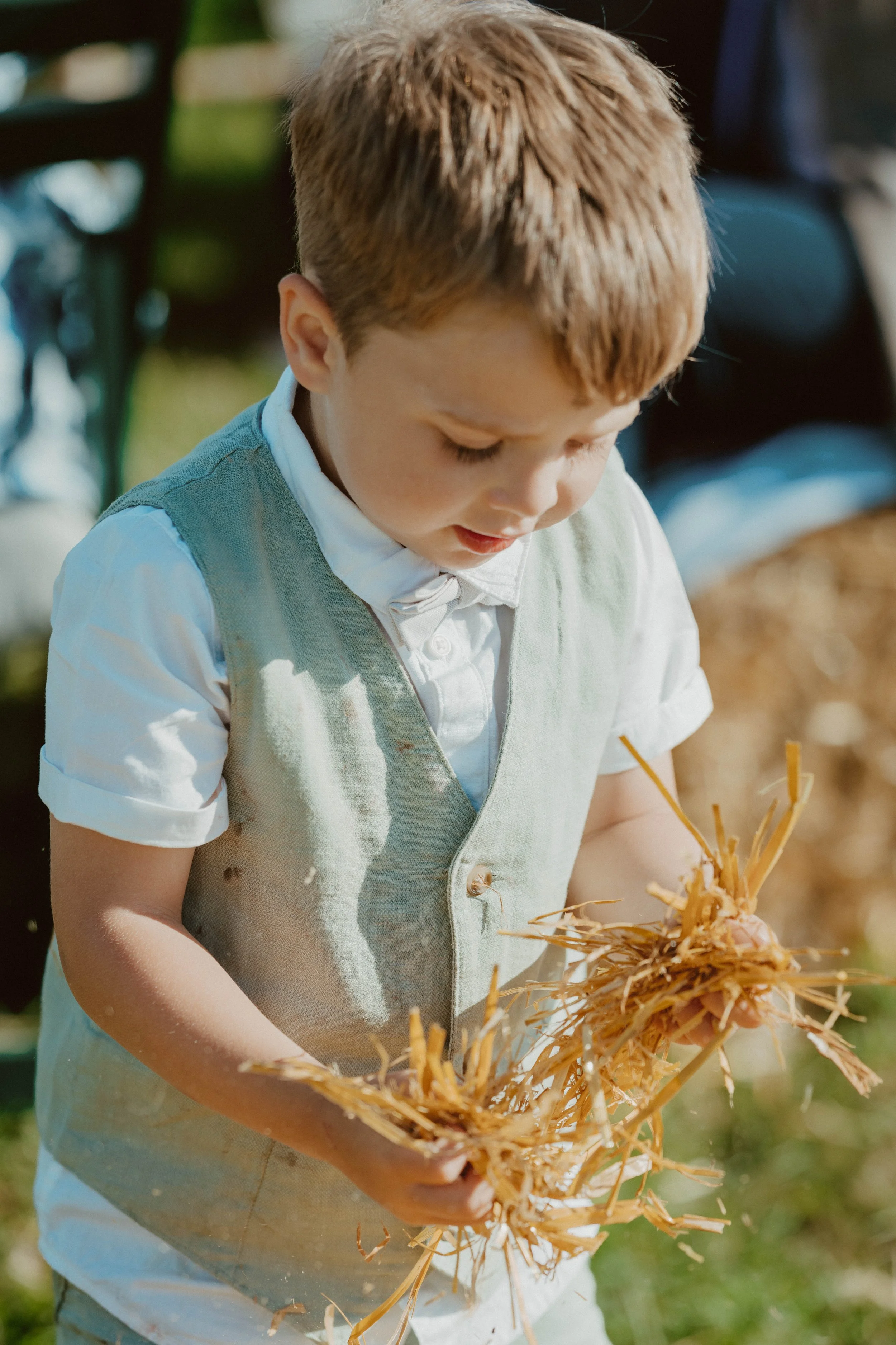 A young boy dressed in vintage clothing holding a bundle of straw or wheat outdoors in a natural setting.