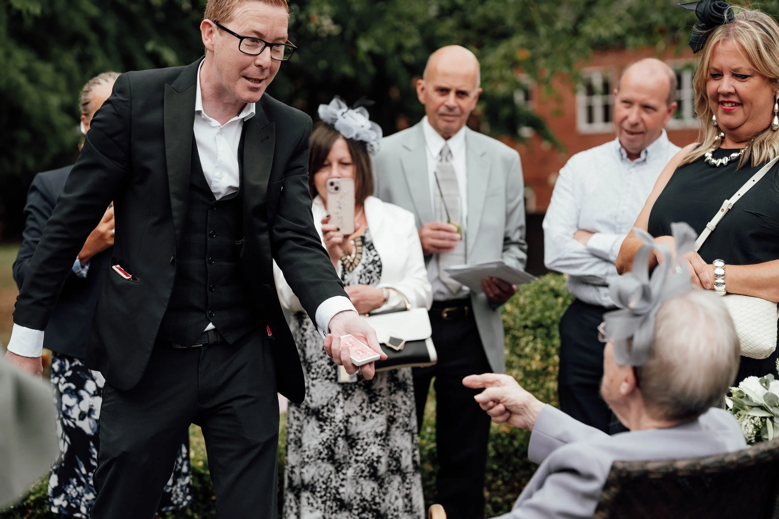A man in a black suit performs a magic trick for an elderly woman with white hair outdoors at a social gathering, surrounded by people in formal attire.