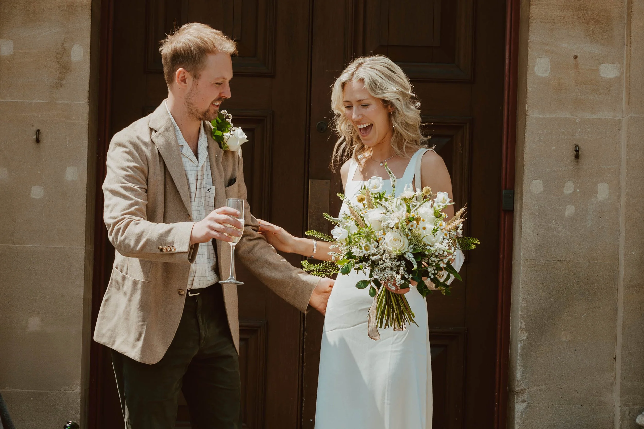 A bride and groom sharing a joyful moment on their wedding day. The bride is holding a large bouquet of white and pink flowers, and the groom is holding a glass of champagne, gesturing towards her.