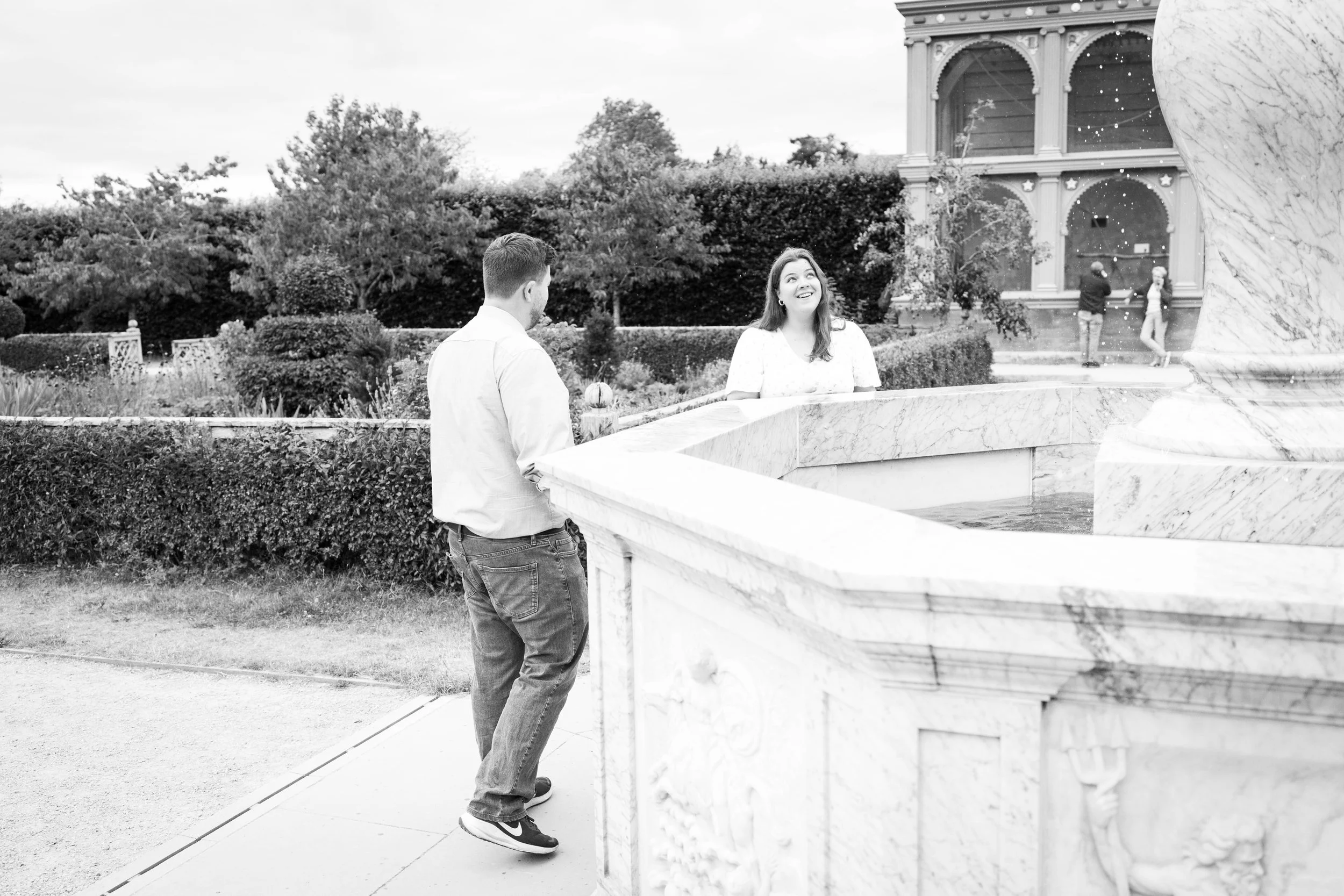 A man and woman stand near a large marble fountain in a garden, with the woman smiling and looking up, while a child and another person are in the background near a building.