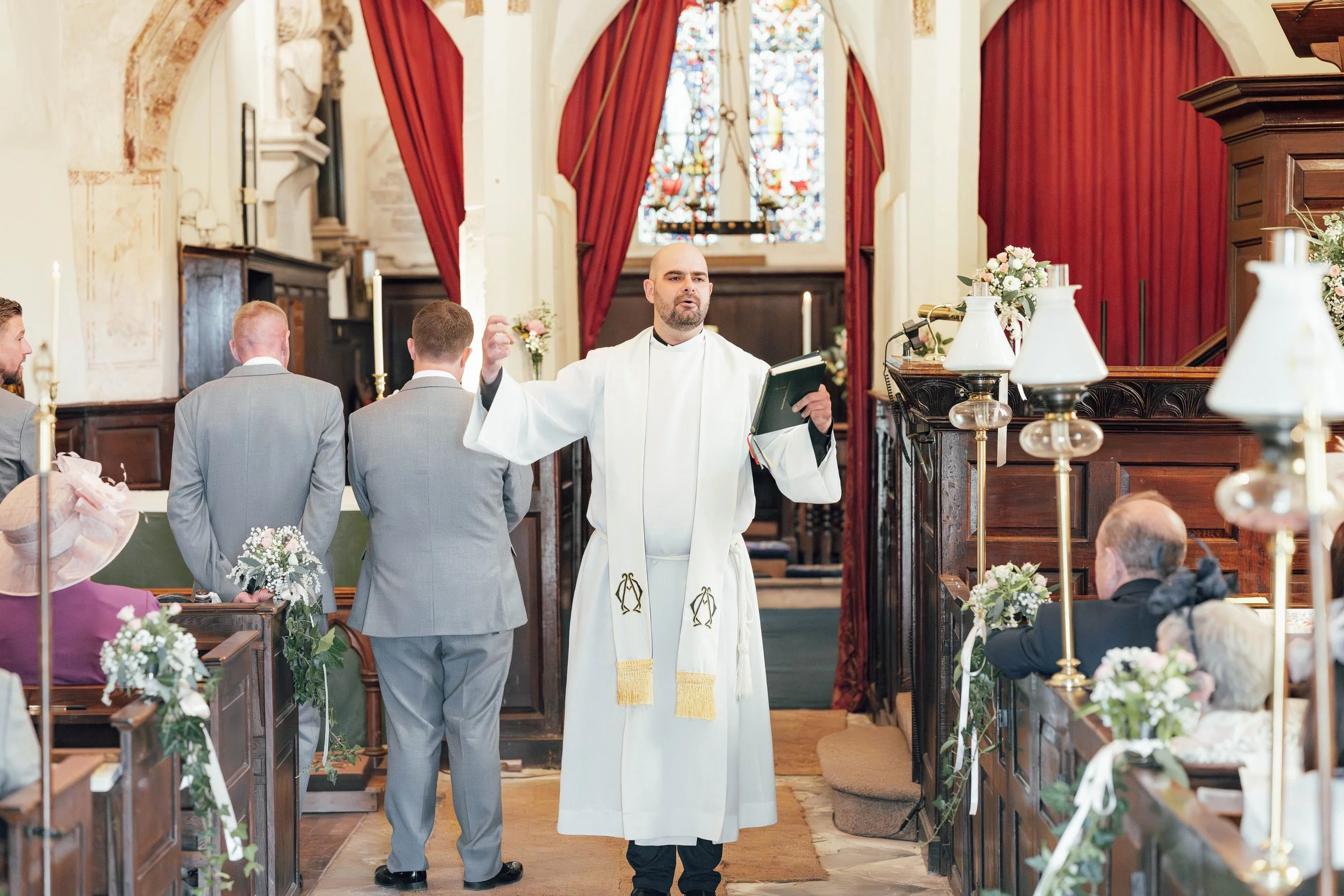 A priest leading a wedding ceremony in a church with groomsmen standing in front of him, decorated with flowers and candles, stained glass windows in the background.