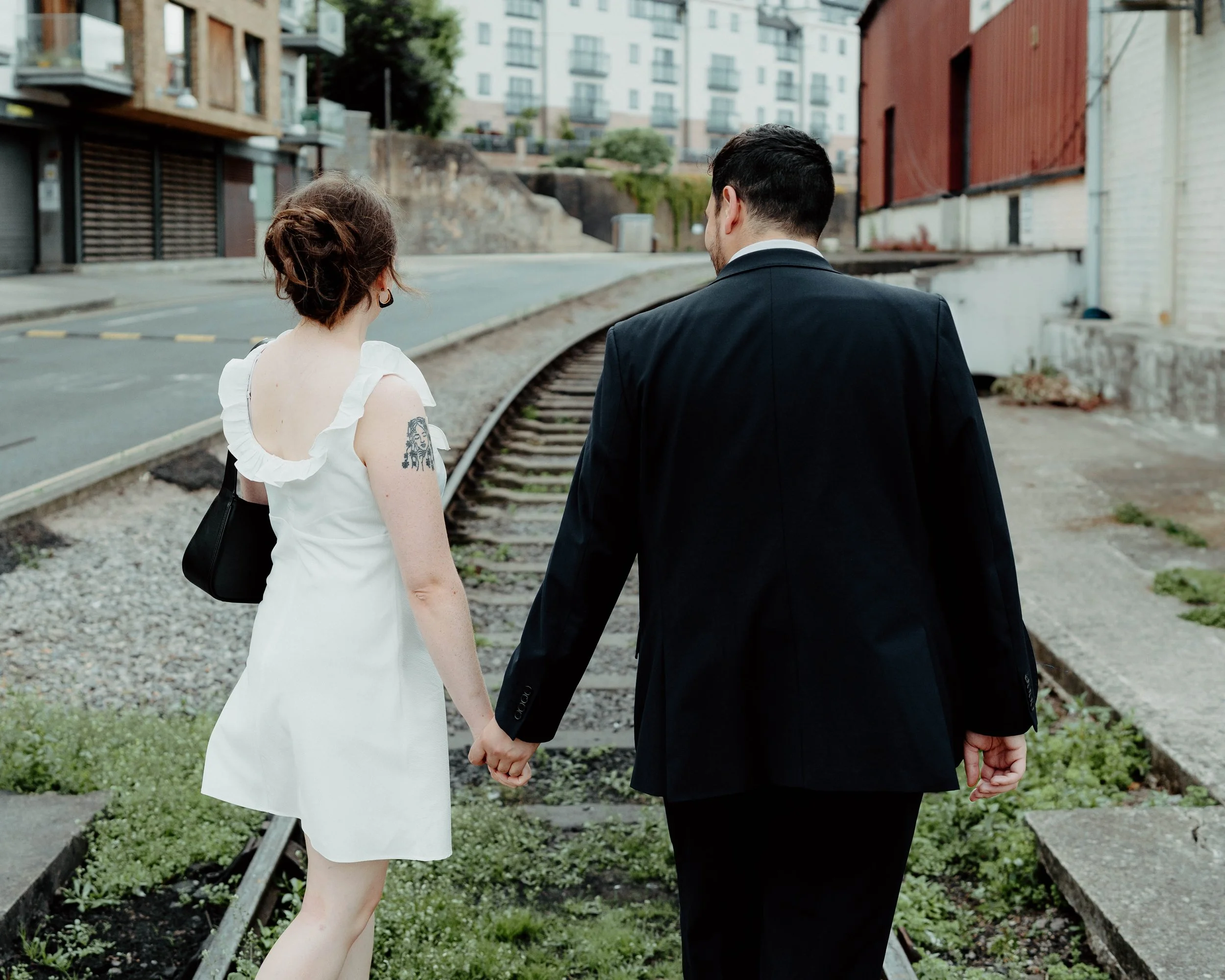 A woman in a white dress with a tattoo on her right arm and earrings, holding hands with a man in a black suit, walking along a railway track in an urban area.
