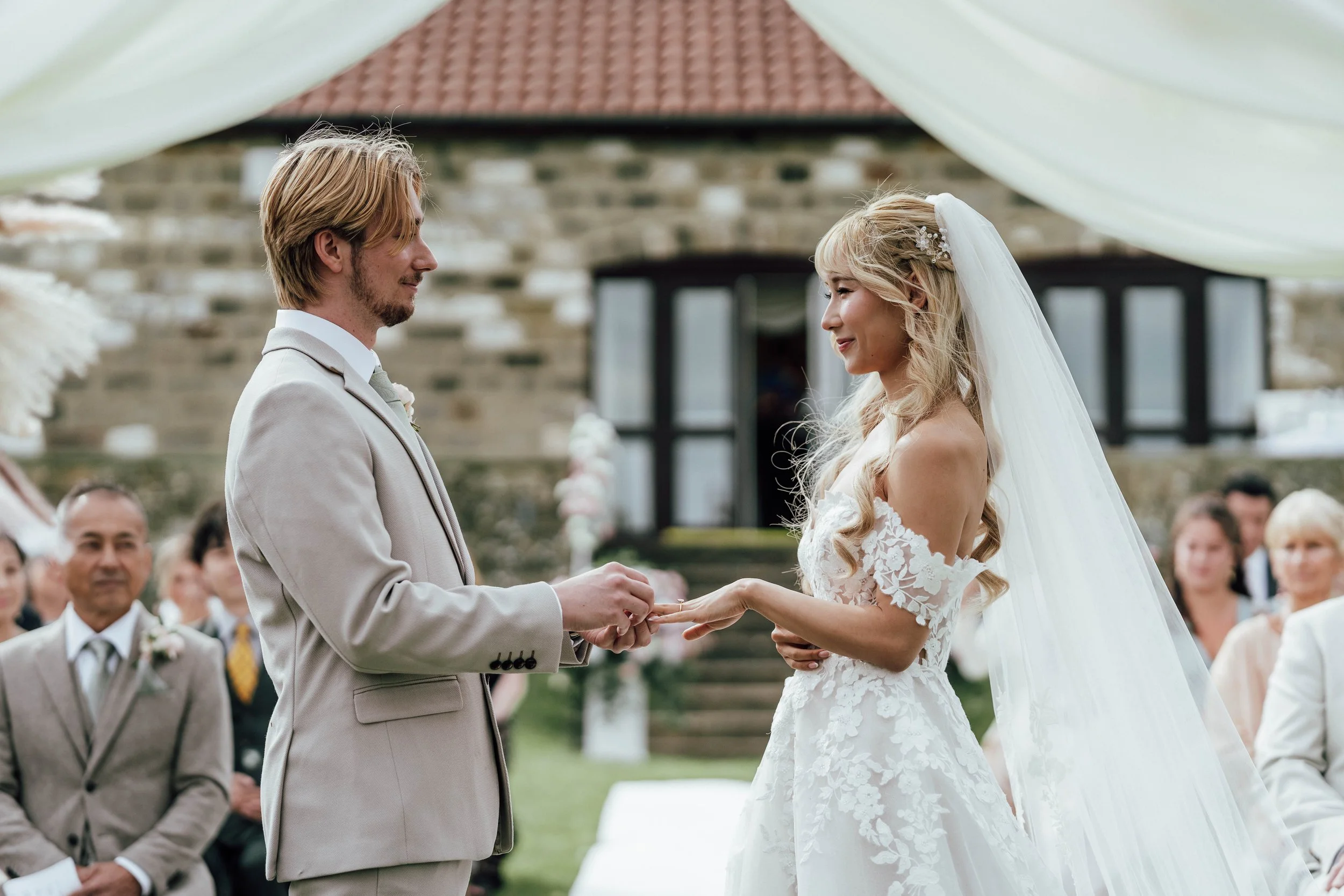 Bride and groom exchanging rings during an outdoor wedding ceremony, with guests seated behind them.