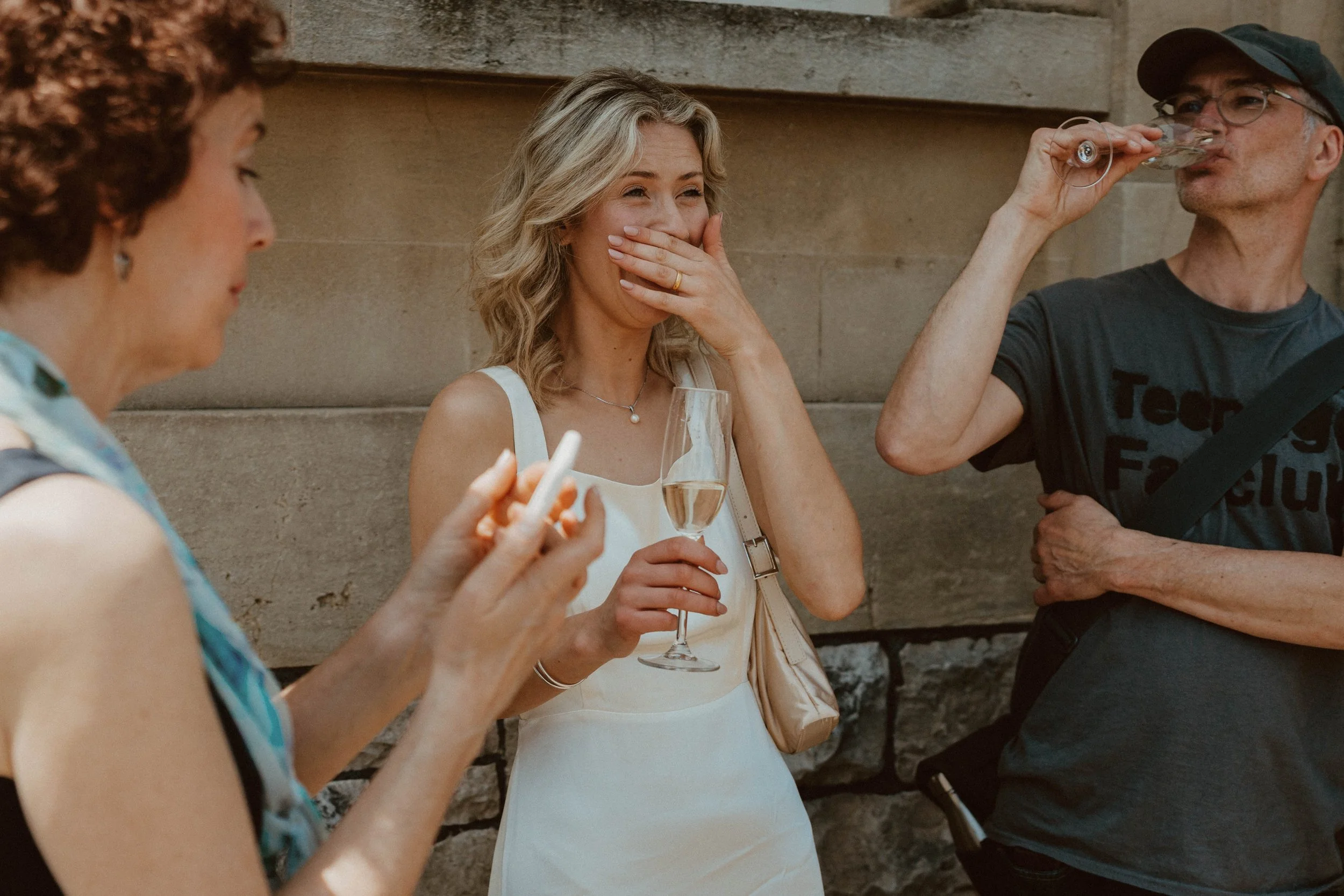Three people standing outdoors against a stone wall, with the woman in the middle holding a glass of champagne and laughing as she covers her mouth, another woman on the left using her phone, and a man on the right drinking from a glass, wearing a ca