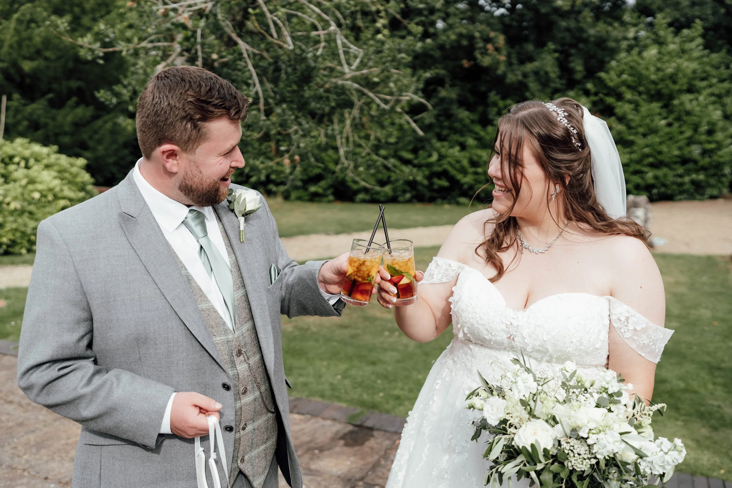 A bride and groom in wedding attire toasting with drinks outdoors, smiling at each other with a green, lush background.