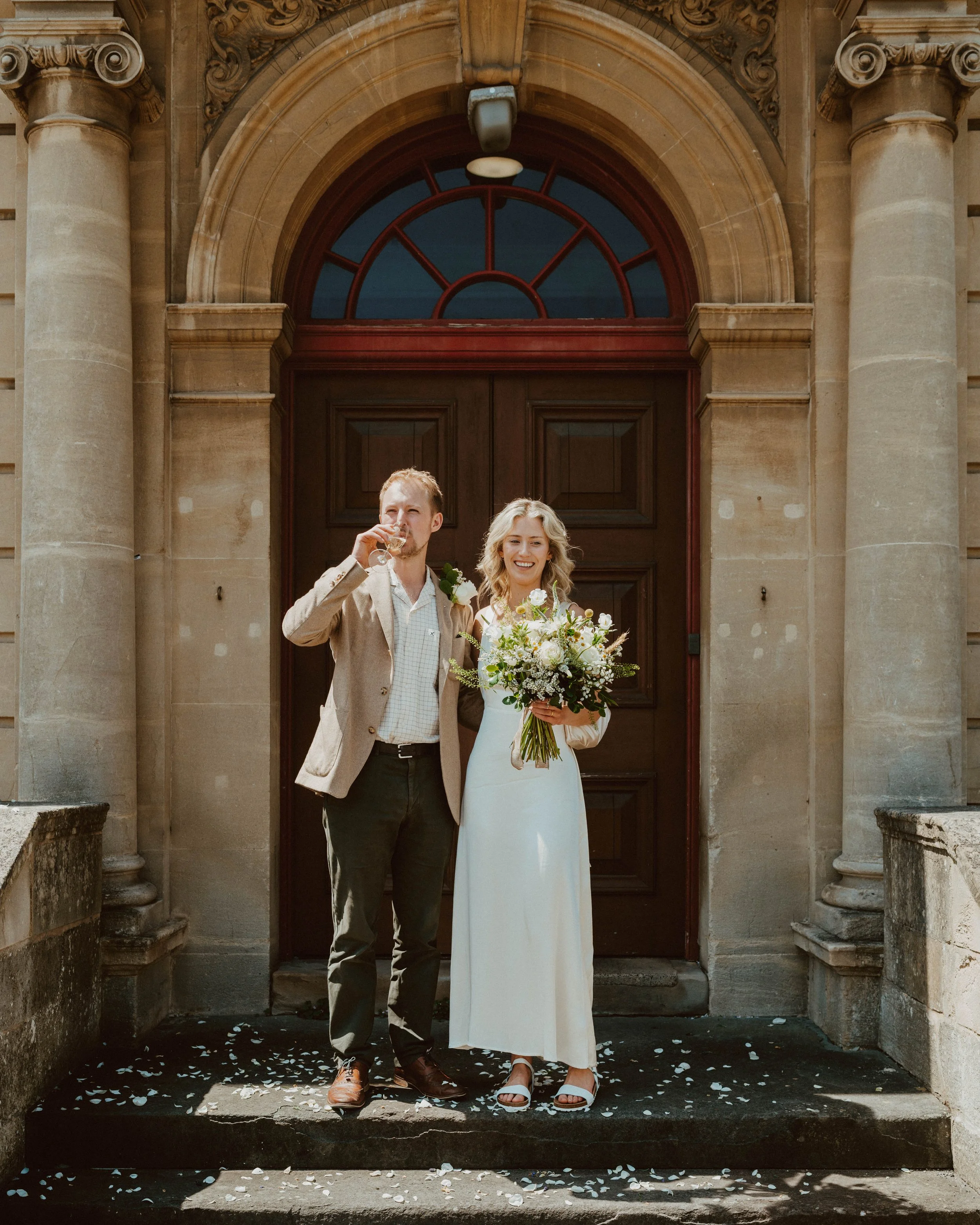 A newlywed couple stands on the steps in front of a historic building with large wooden doors and stone columns. The bride is holding a bouquet of white flowers and smiling, while the groom is drinking from a glass. Flower petals are scattered on the