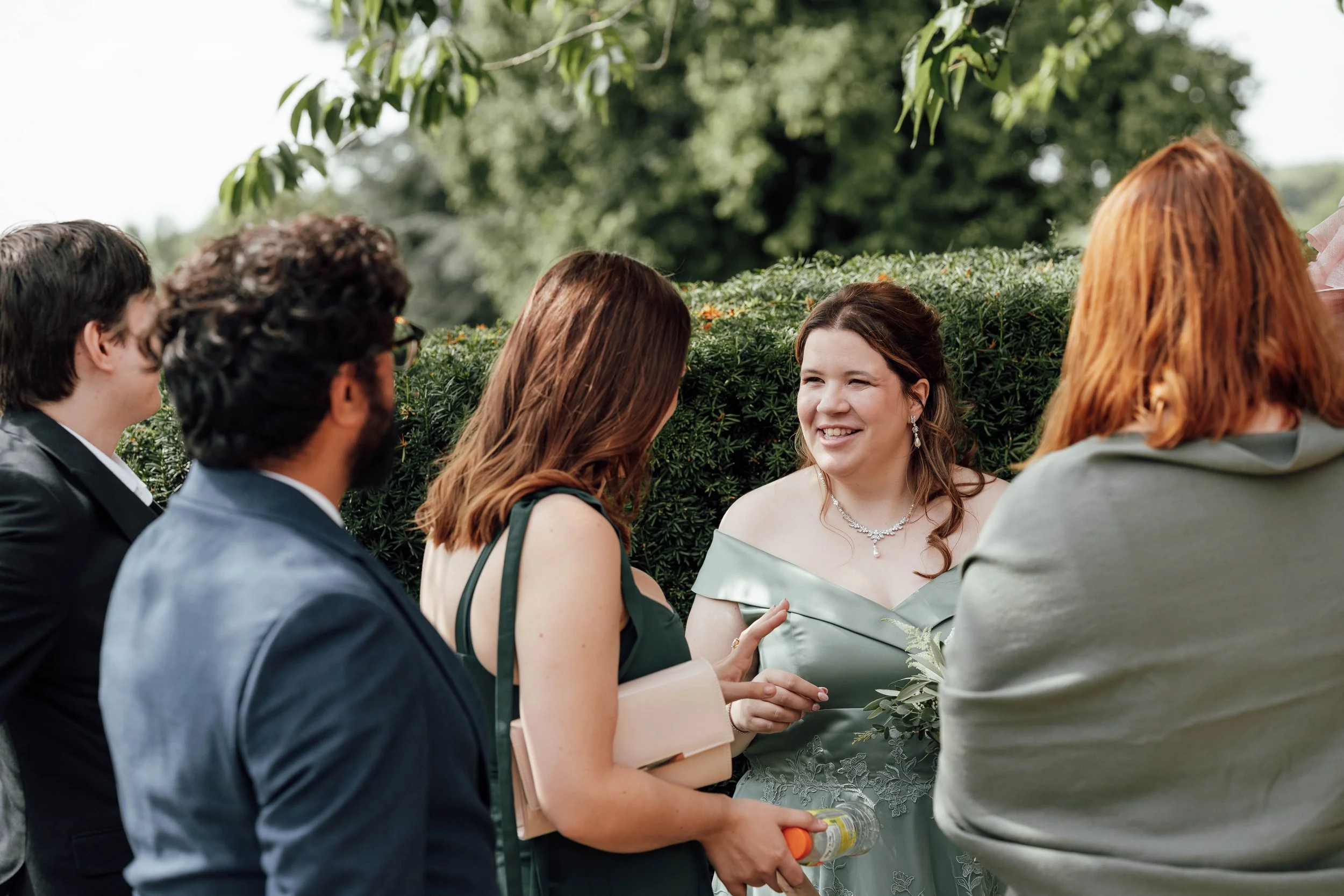 Group of people talking outdoors at a garden wedding. A woman in an off-shoulder gown is smiling as she talks with others.
