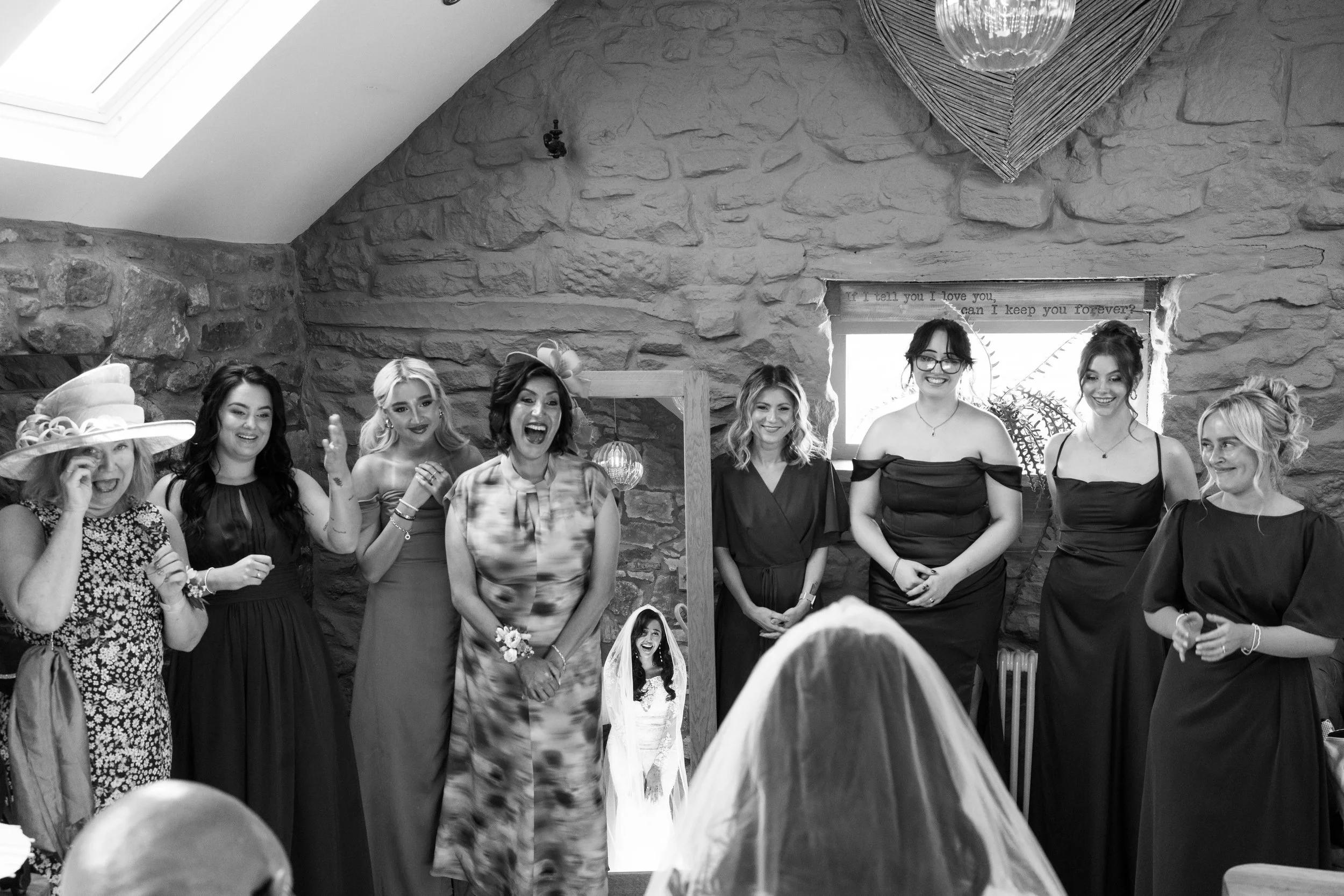 Group of women at a wedding reception, with one woman in a white dress and veil, possibly the bride, standing in the middle. They are standing against a stone wall with a small window. Some women are smiling, laughing, and looking at the bride.