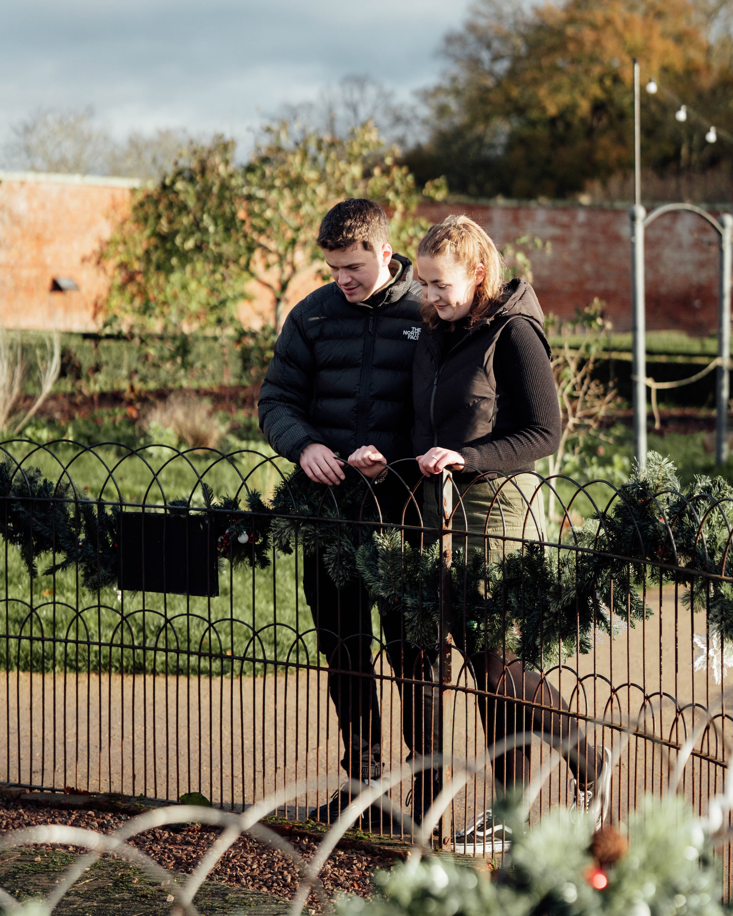A young man and woman looking at Christmas wreaths on a park fence during daytime.