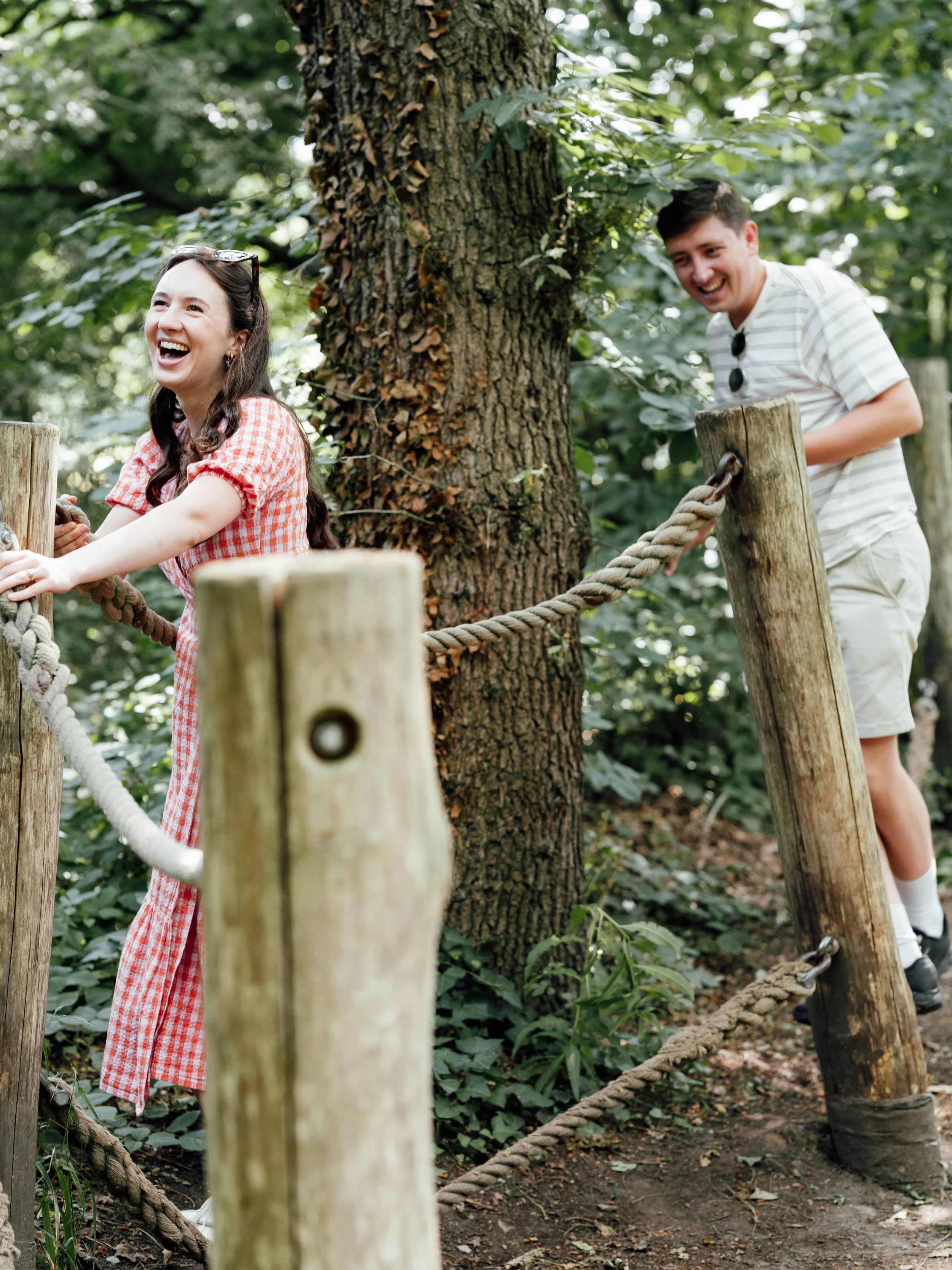 A joyful woman in a red and white checkered dress leaning on a rope fence, with a man in a striped white shirt and shorts behind her, both in a lush green outdoor forest.