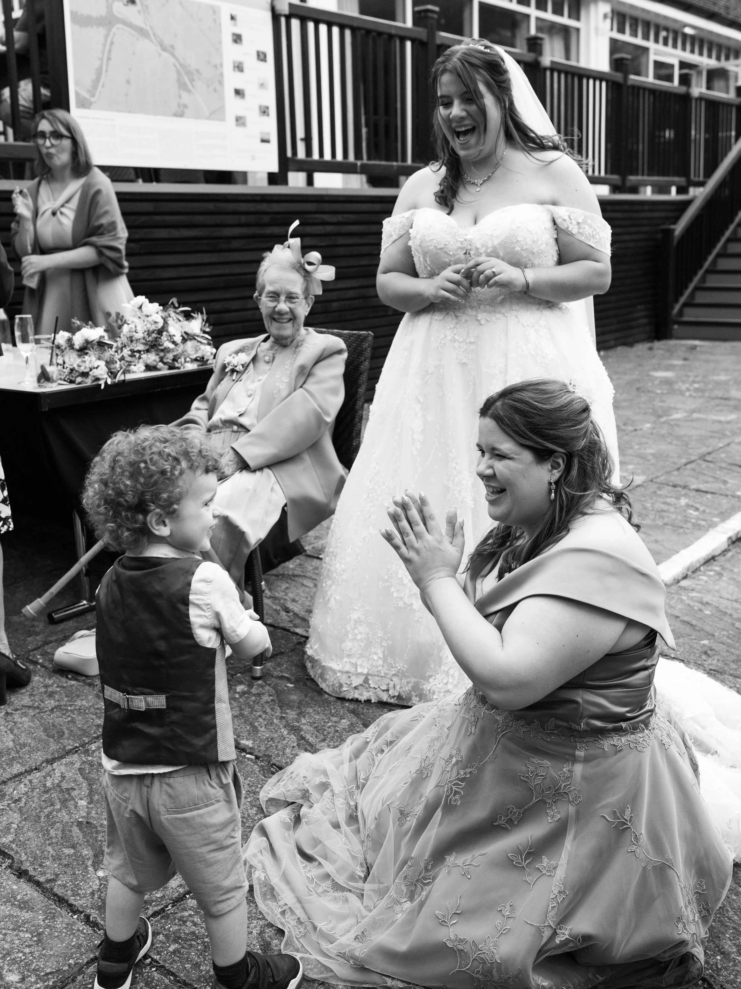 A wedding celebration taking place outdoors with a bride in a strapless gown, a woman kneeling in a fancy dress, an elderly woman in glasses and a blazer sitting at a table with flowers, a young boy with curly hair and a vest standing, and another wo