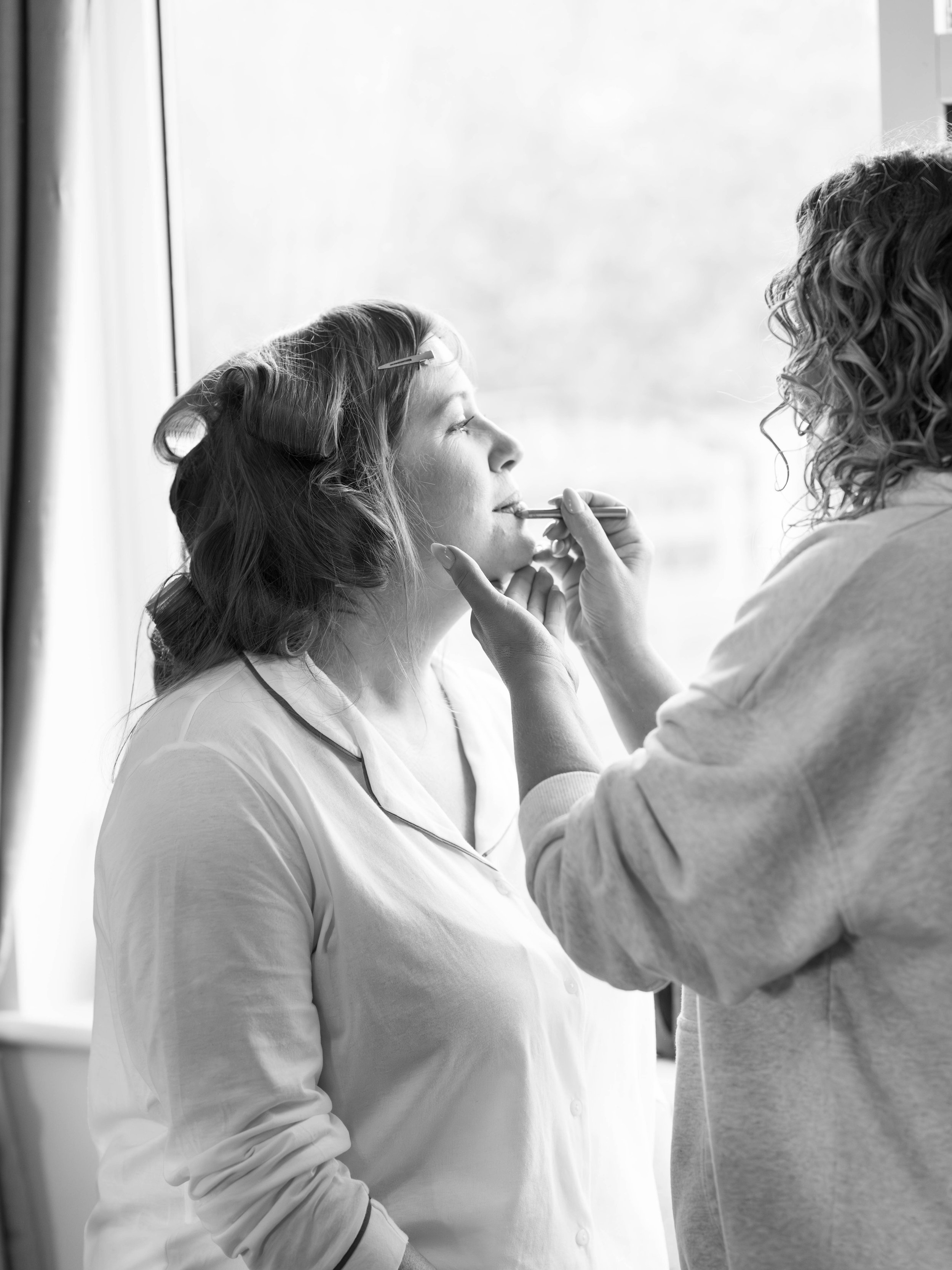 A woman getting her lipstick applied by a makeup artist in front of a window.