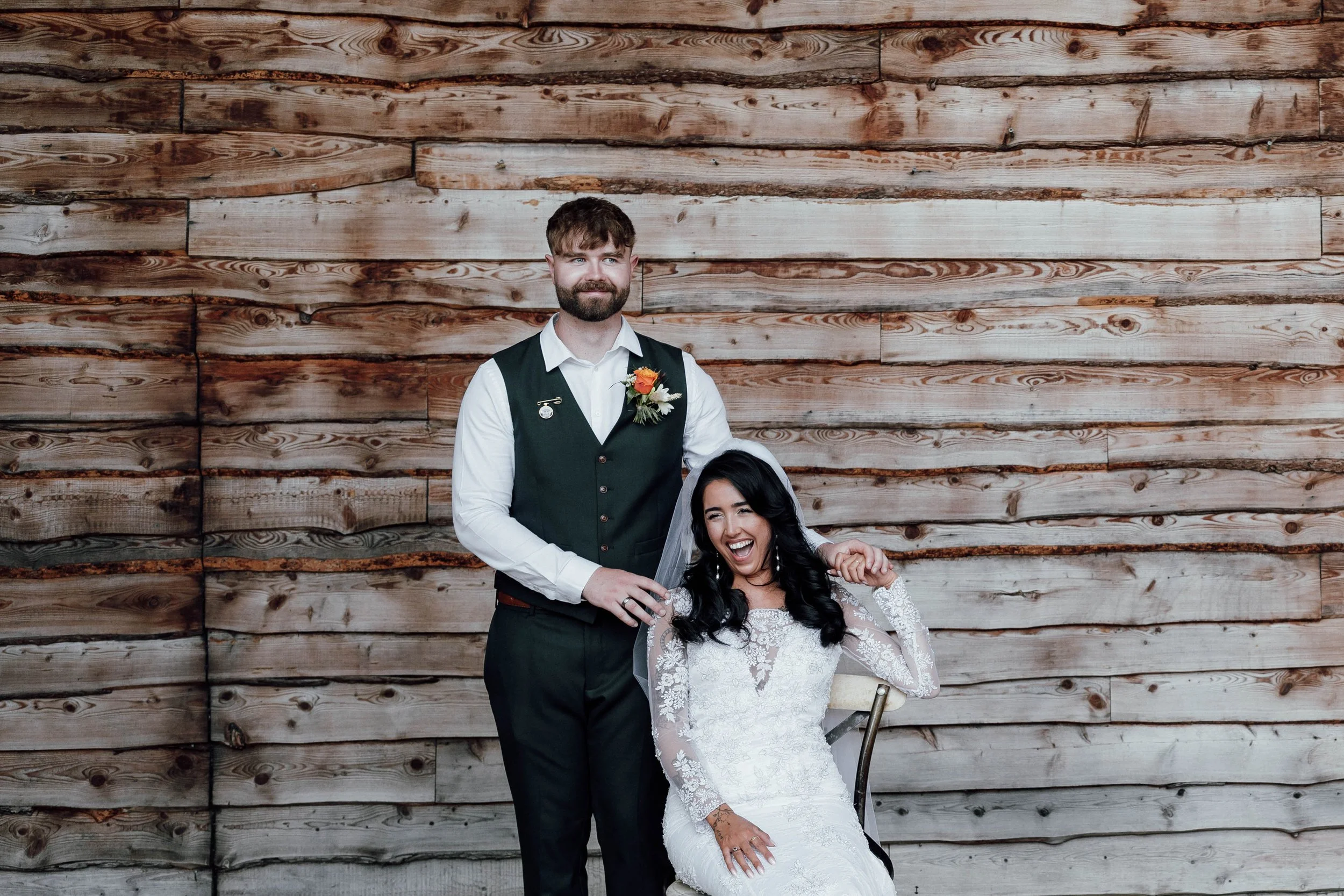 A smiling bride in a white lace wedding dress and veil, sitting on a chair, with a groom standing next to her, against a rustic wooden wall background.