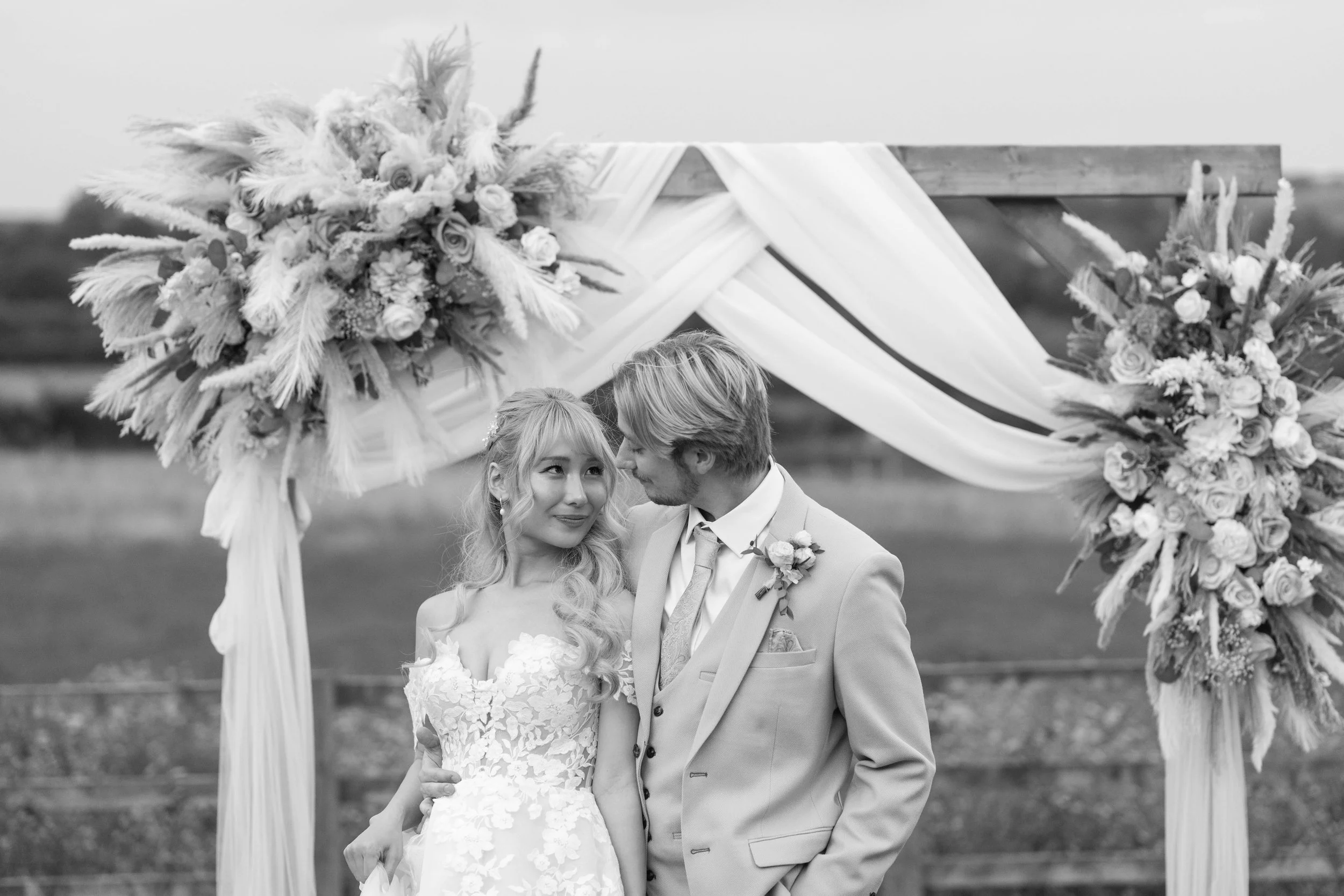 A wedding couple standing under an arch decorated with flowers and draped fabric, outdoor setting, black and white photography.