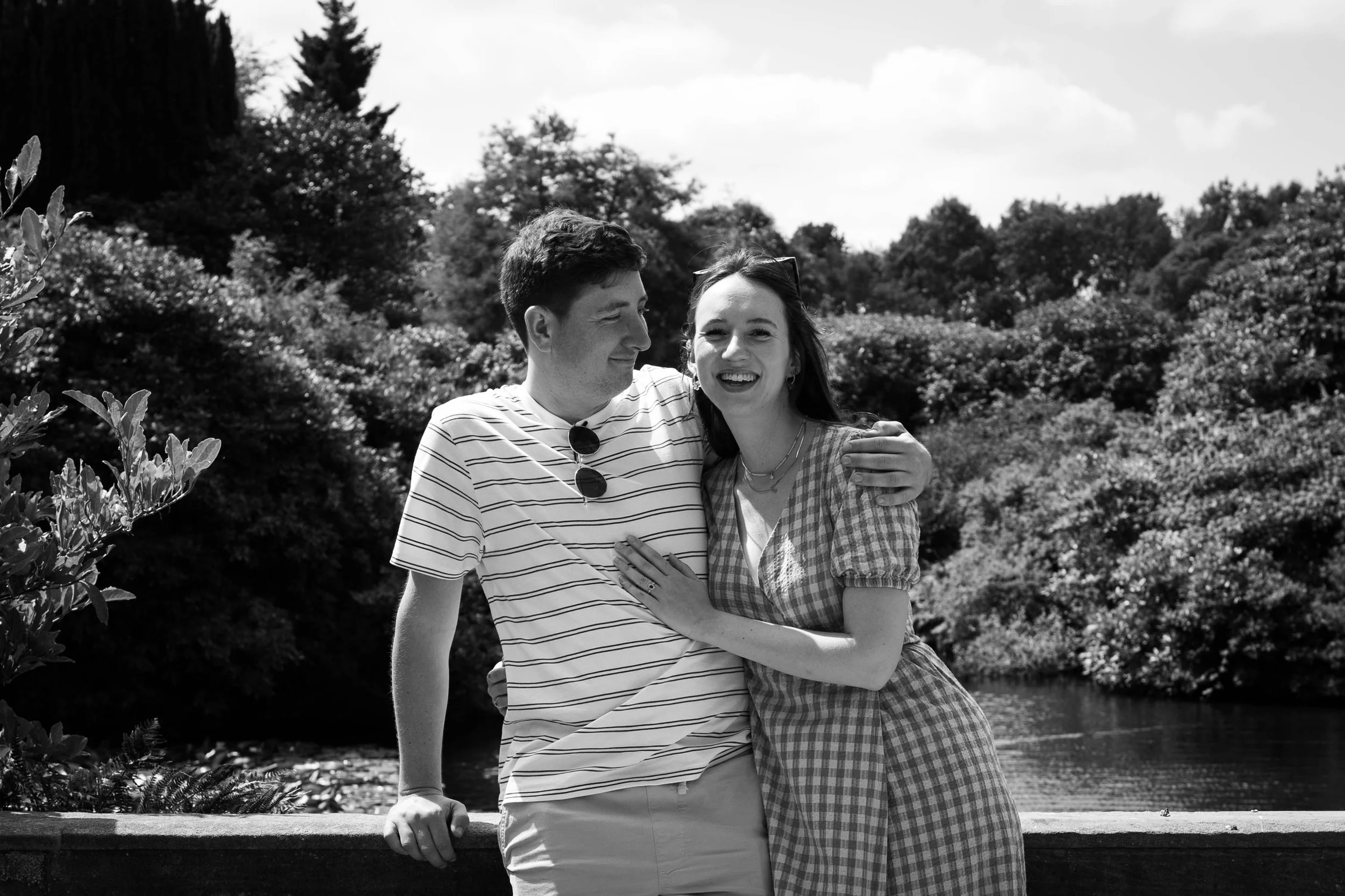 A black and white photo of a young couple smiling and hugging outdoors near a body of water, with trees and sky in the background.