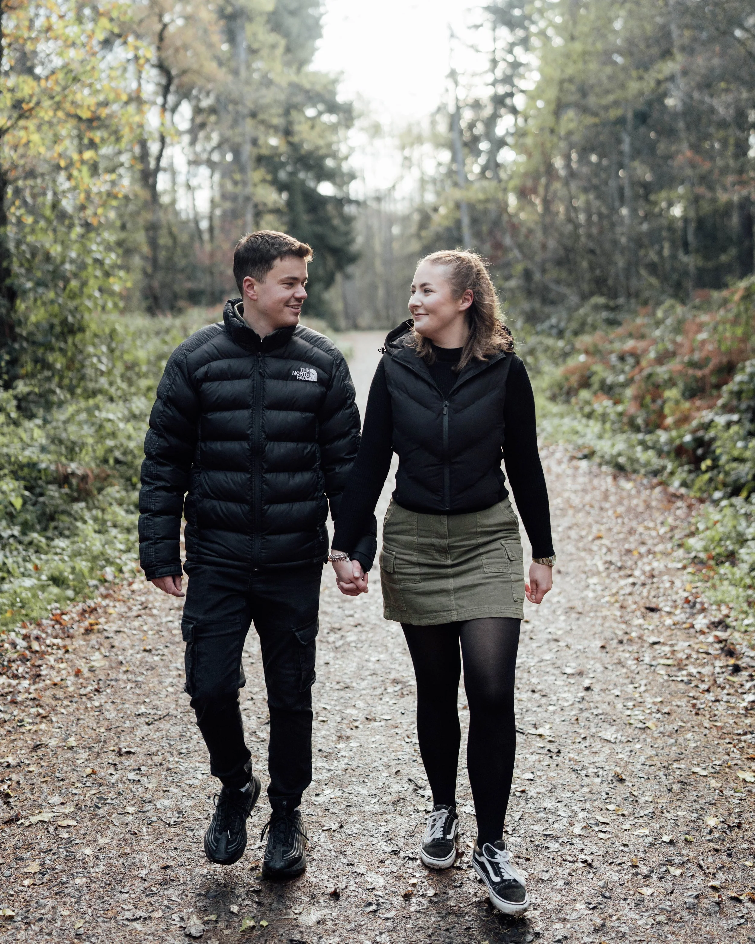 A young man and woman walk hand-in-hand down a wooded dirt trail, enjoying a moment together outdoors.