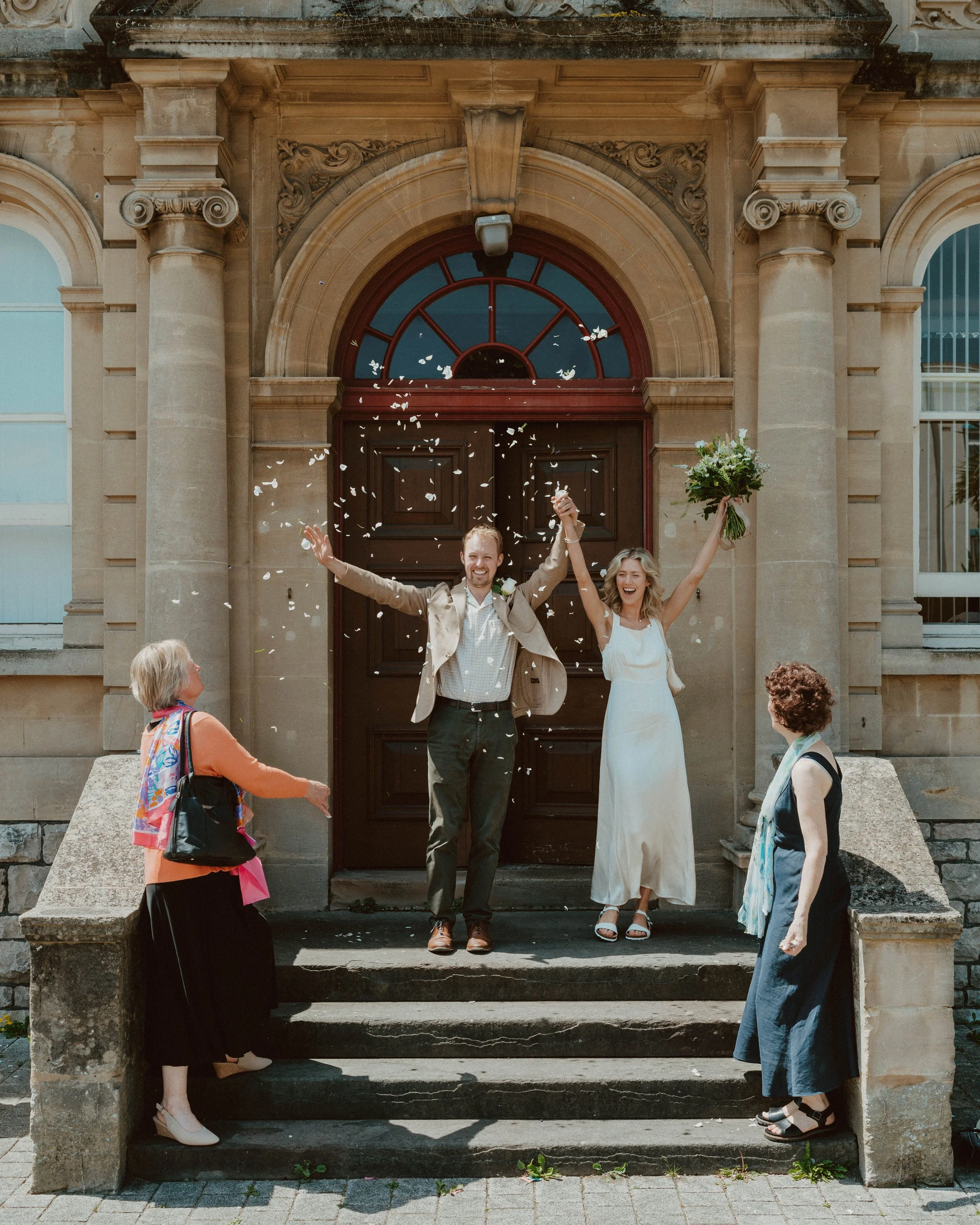 A newlywed couple celebrating on the steps of a building with three older women, with white confetti in the air and the bride holding a bouquet.