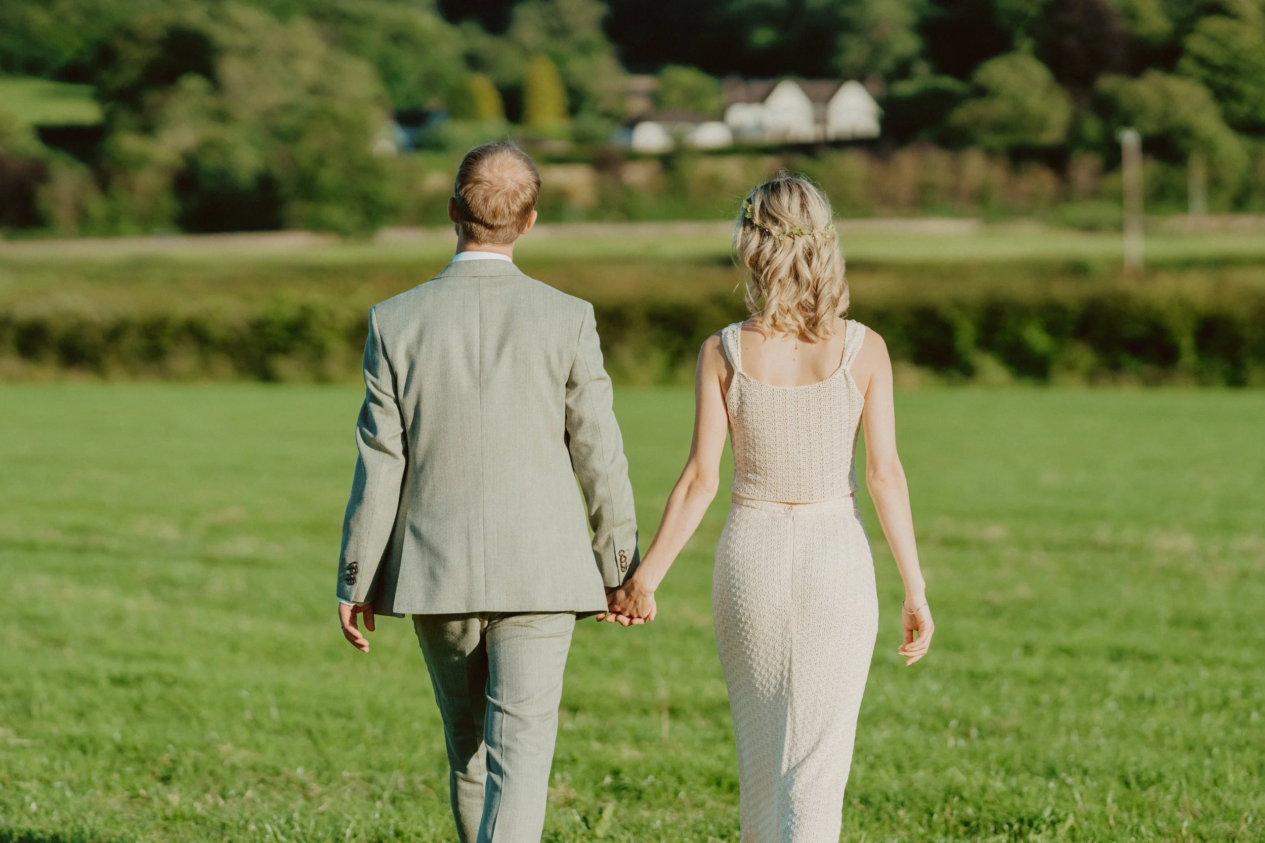 A man and a woman holding hands, walking across a grassy field on a sunny day, with trees and buildings in the background.