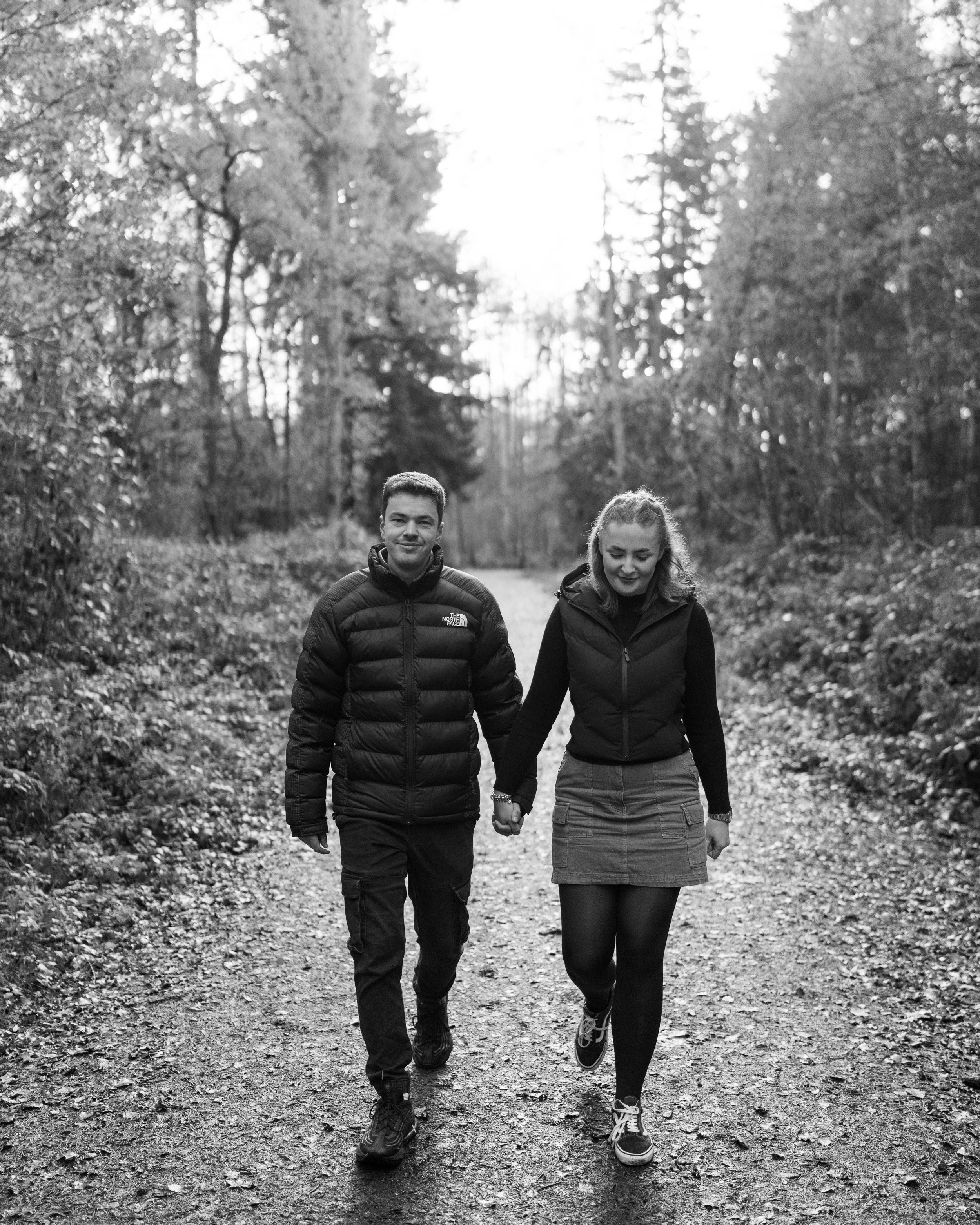A young couple walking hand in hand on a dirt trail in a wooded area during daytime.