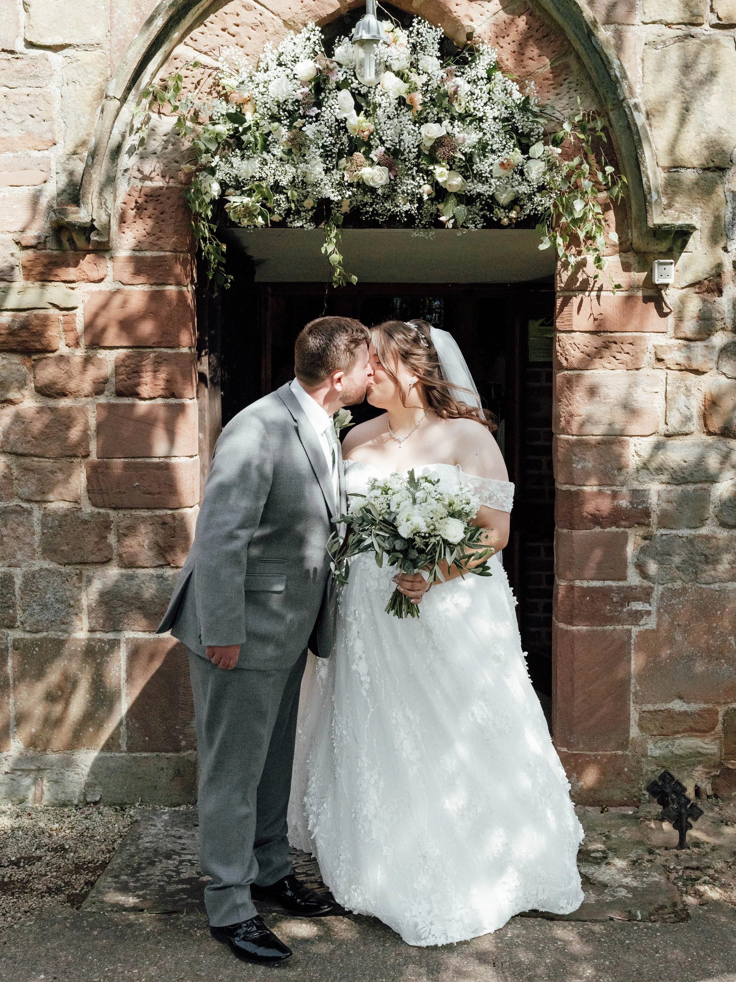 A bride and groom kissing in front of a stone chapel decorated with white and pink flowers.