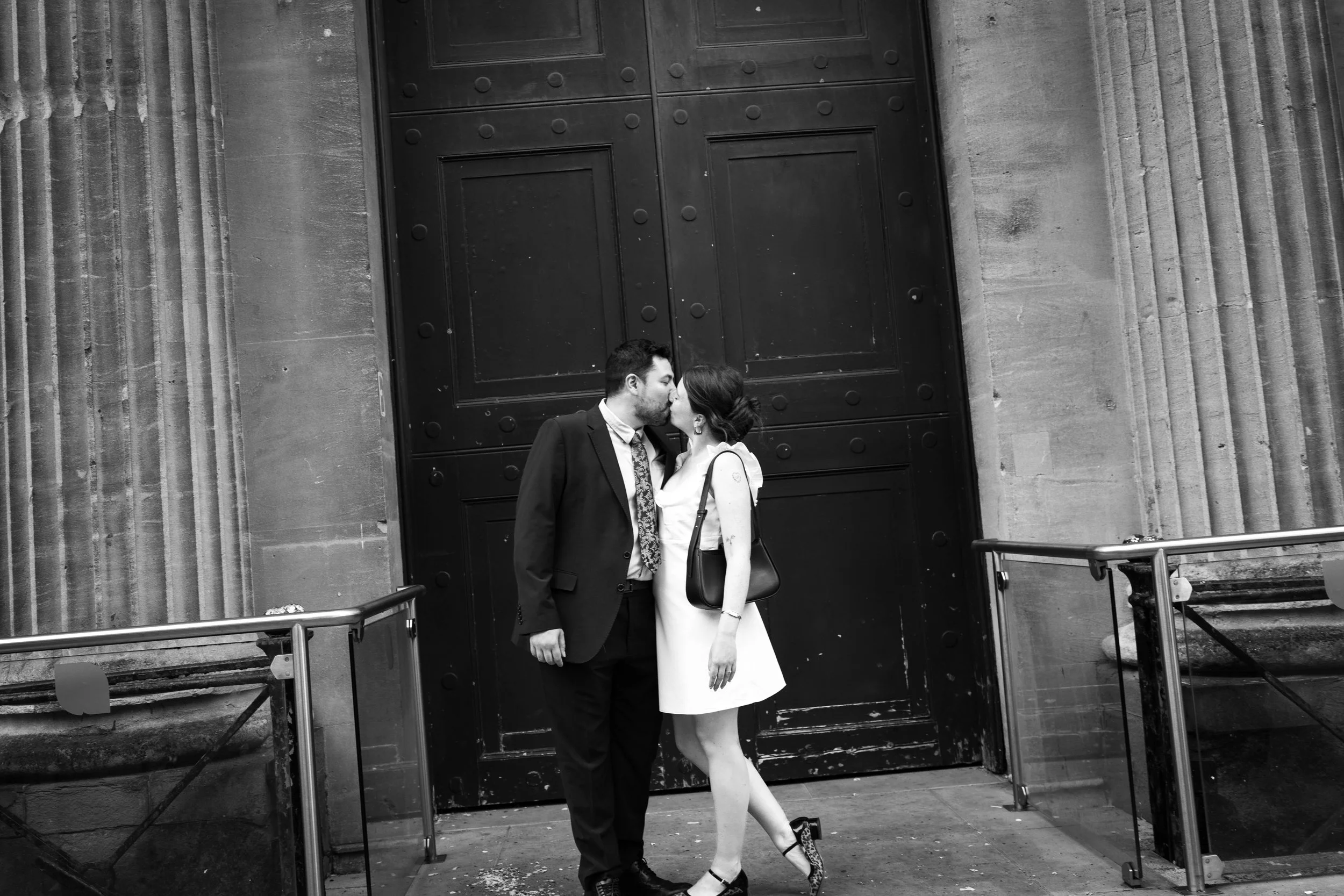 A black-and-white photo of a couple kissing in front of a large black wooden door.