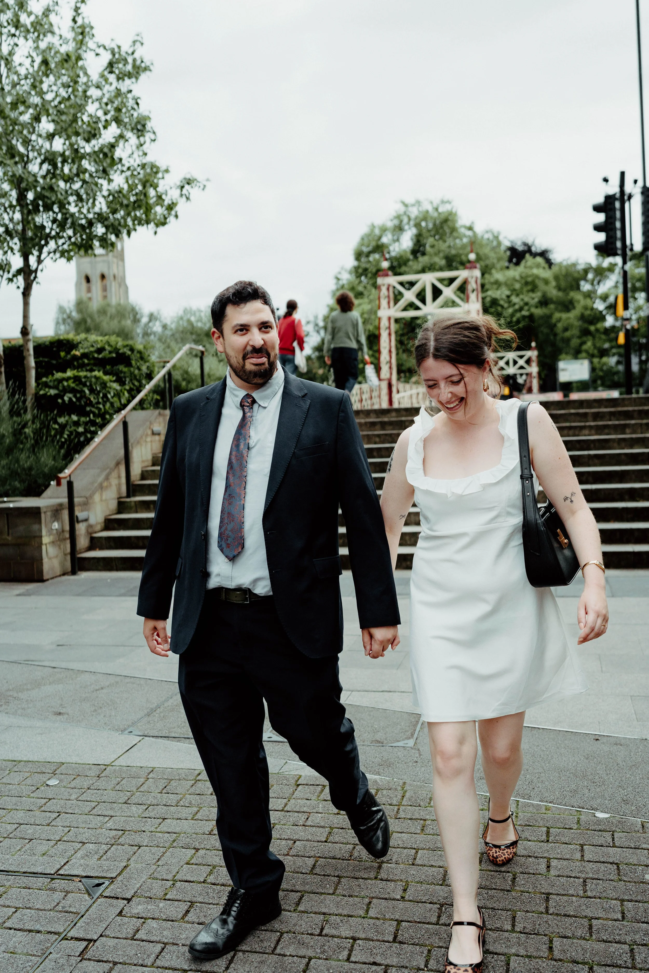 A man and woman holding hands and walking outdoors on a cloudy day, with steps and greenery in the background.