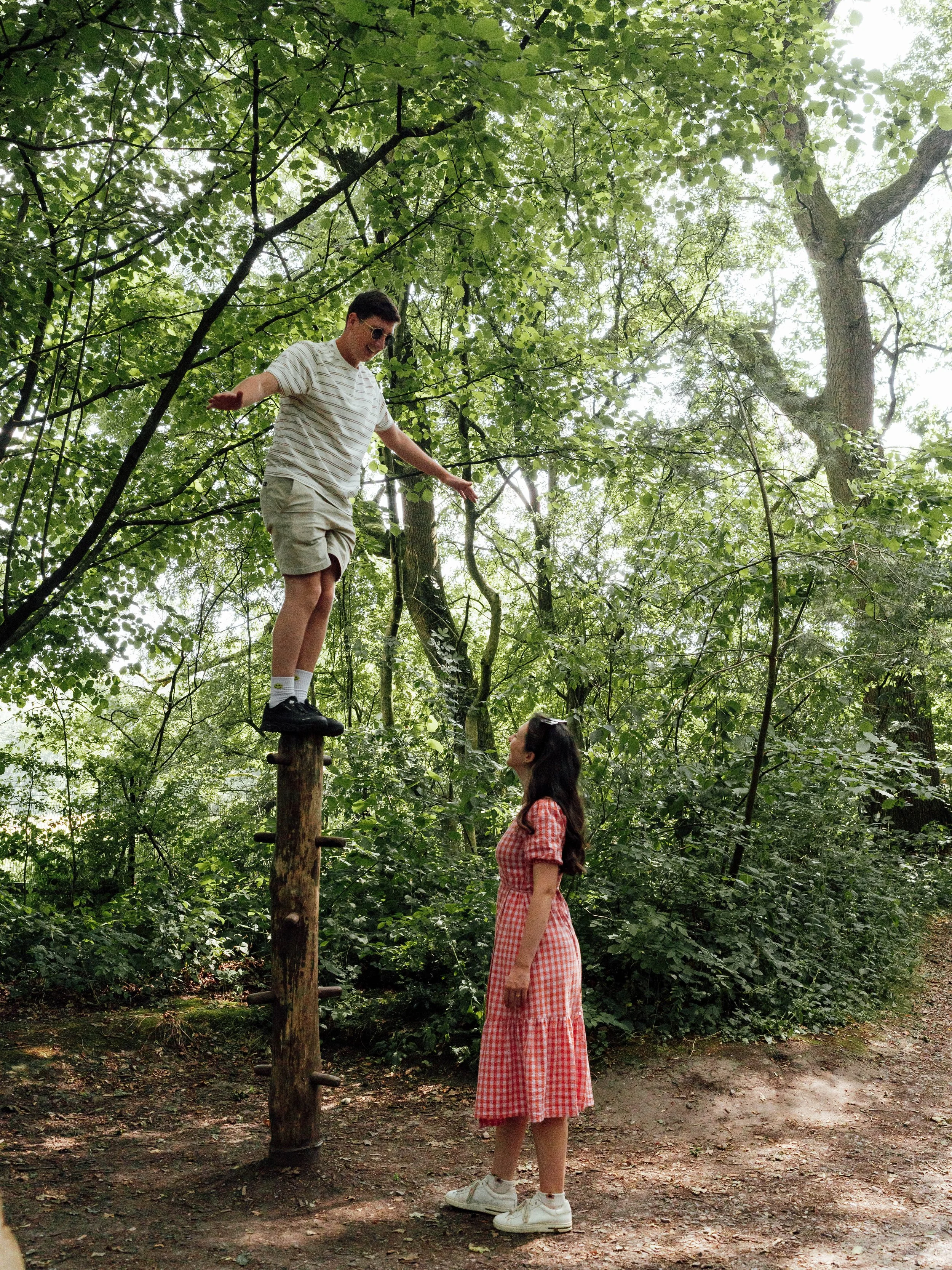 A young man in sunglasses, a striped t-shirt, and beige shorts balancing on a wooden knotted pole in a green forest, with a young woman in a red gingham dress and white sneakers standing on the ground looking up at him.