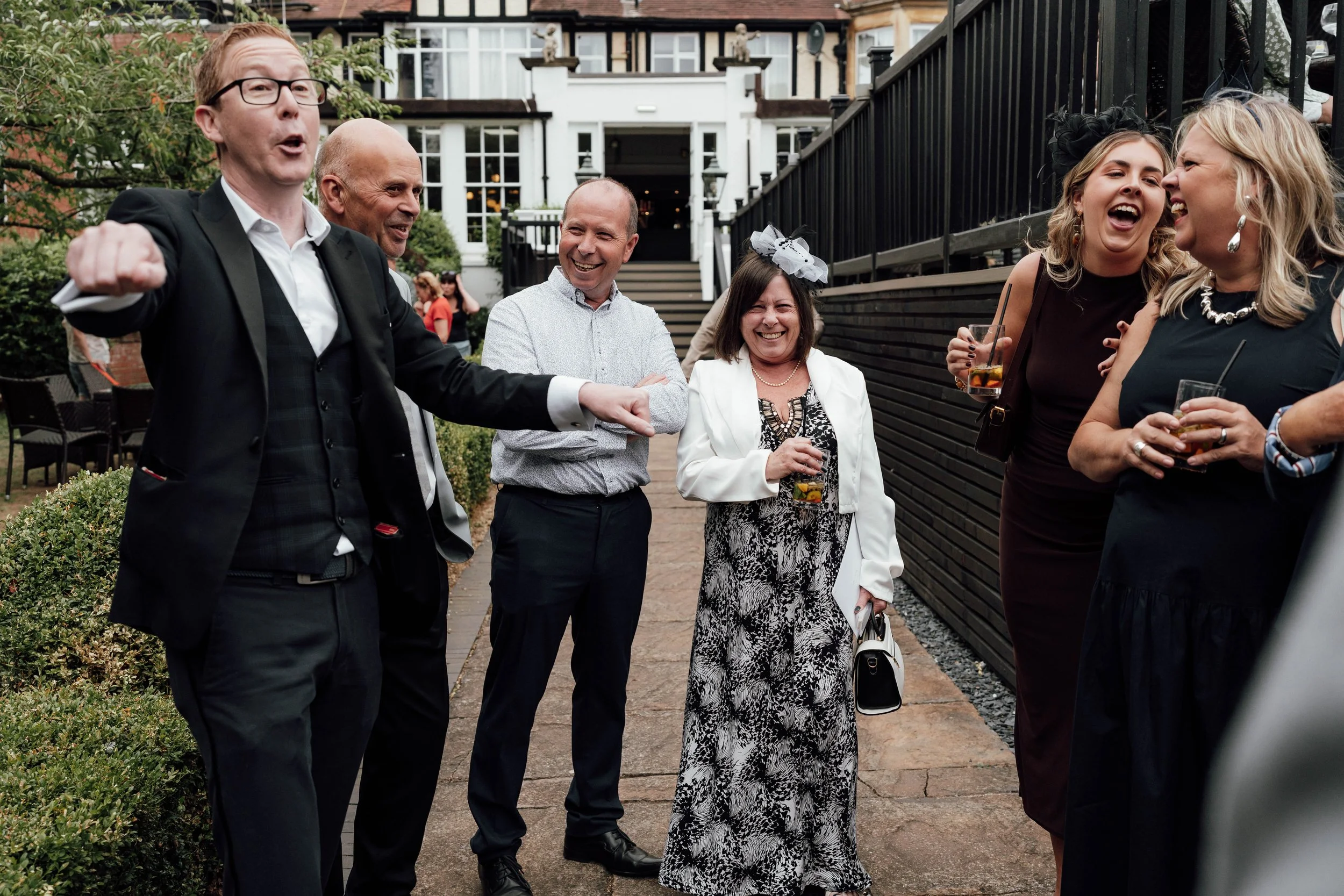 Group of people at an outdoor gathering, smiling and talking, some holding drinks, with a building in the background.