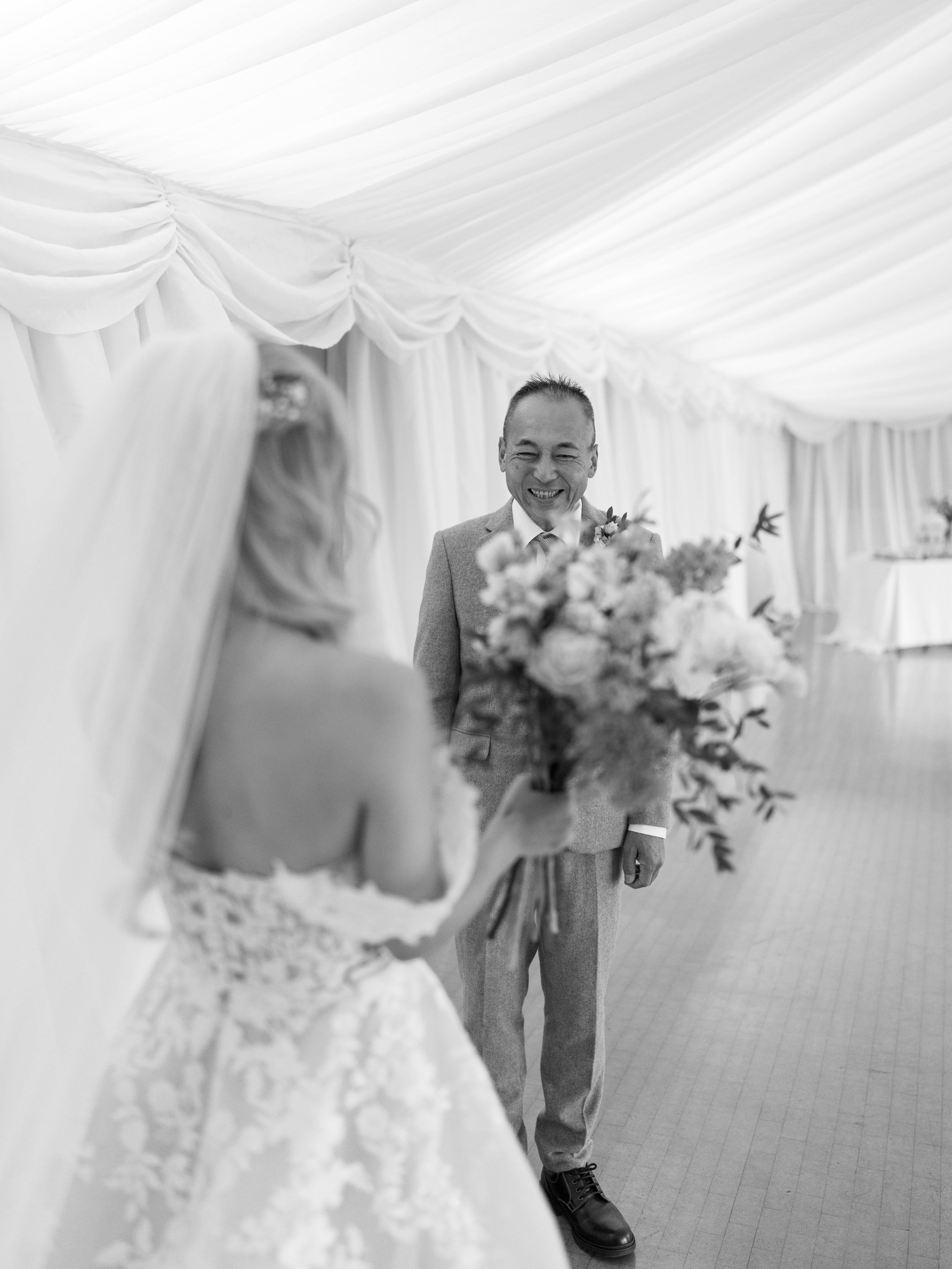 A black-and-white photo of a bride presenting a bouquet to a smiling man, likely her father, at a wedding ceremony inside a decorated tent.