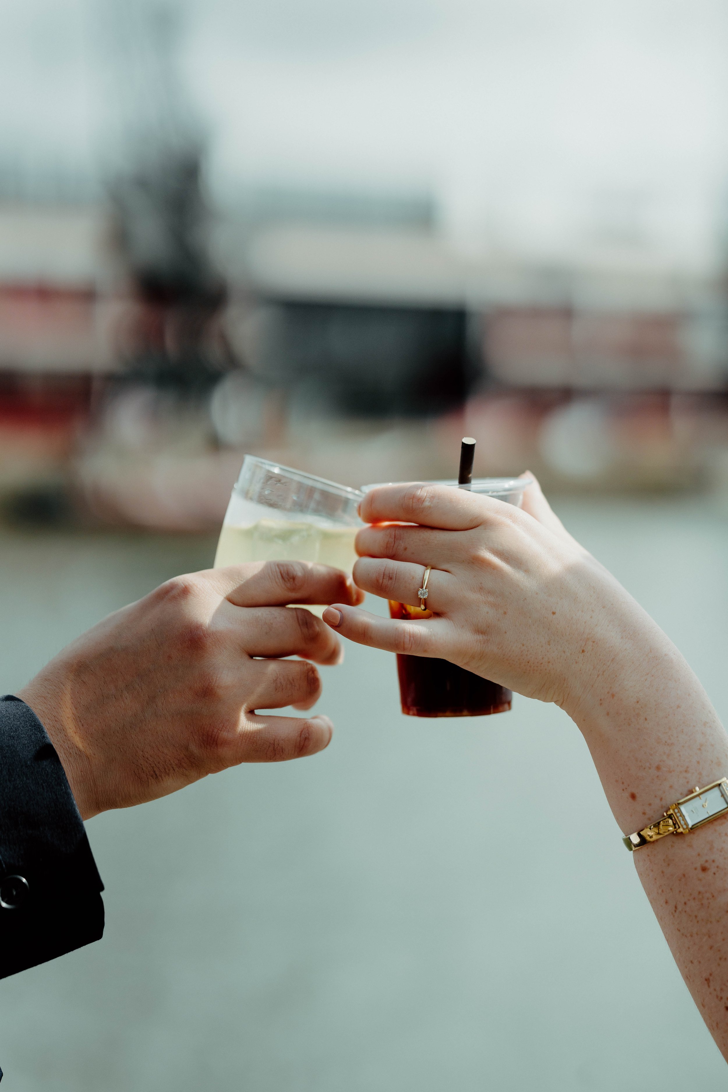 Two hands holding drinks in a toast, one glass of clear beverage, the other of dark soda, with a blurred background.