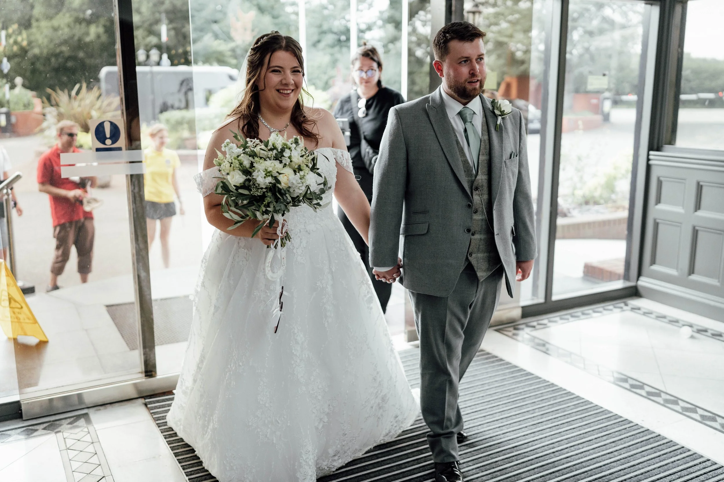 A bride and groom holding hands as they walk inside a building, smiling. The bride is wearing a white wedding dress and holding a bouquet of flowers. The groom is dressed in a gray suit. There are people visible through the glass doors in the backgro
