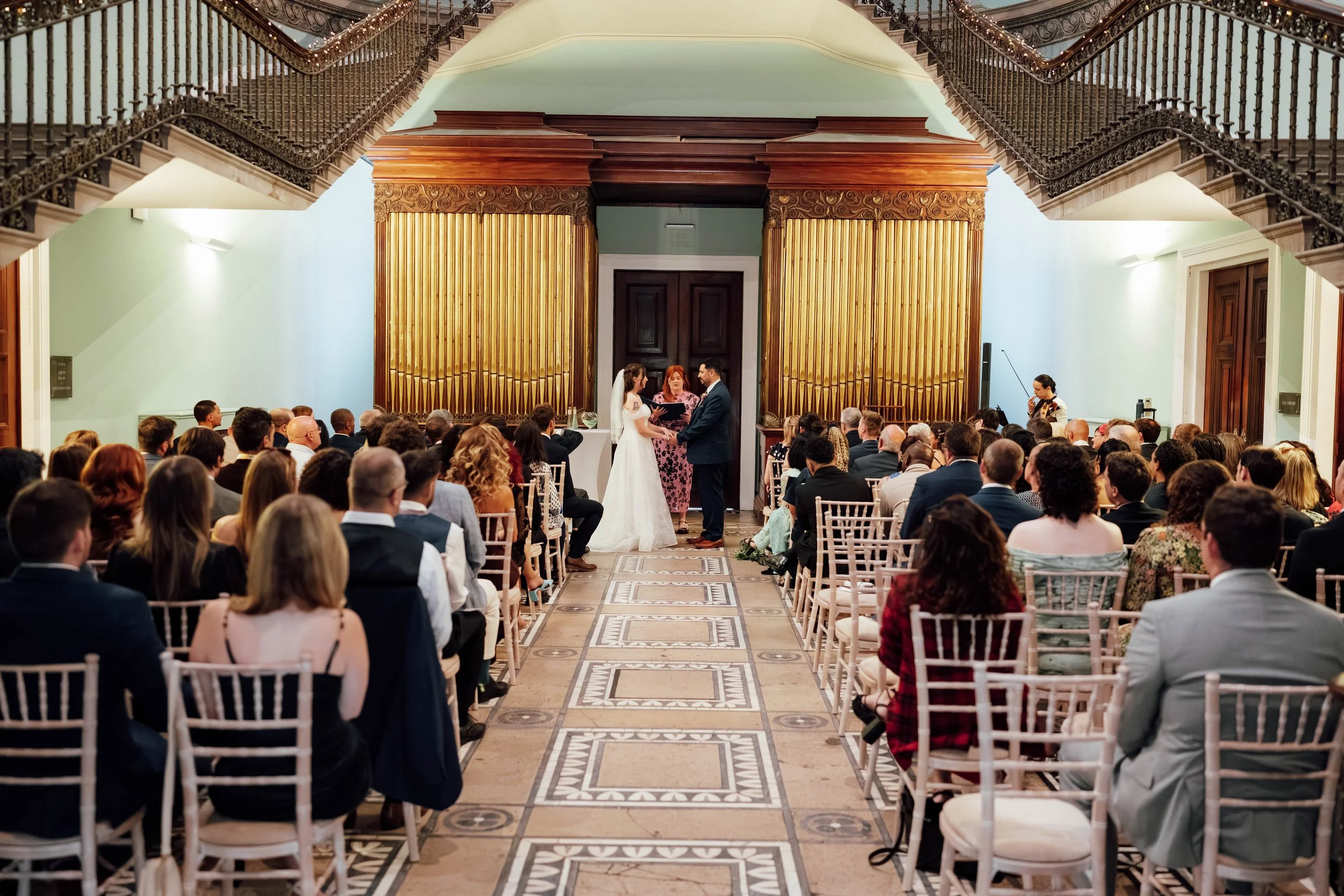 Wedding ceremony with bride and groom exchanging vows in front of an officiant, inside an elegant venue with a patterned tile floor and gold paneling behind the couple, guests seated on either side.