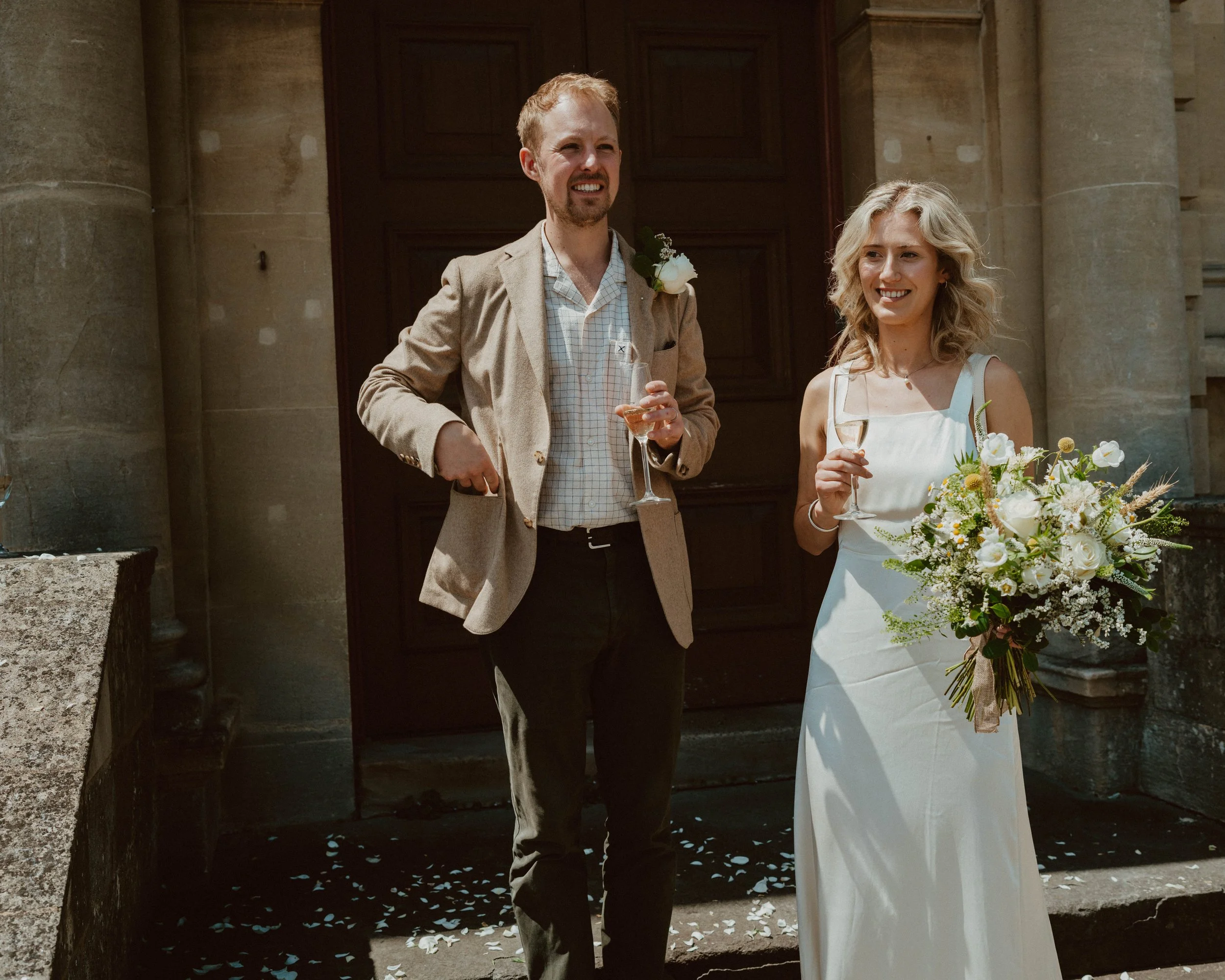 A bride and groom standing outside a church, smiling and holding glasses of champagne. The bride is holding a bouquet of white and green flowers, and the groom is wearing a beige blazer with a boutonniere. There are white flower petals on the steps.