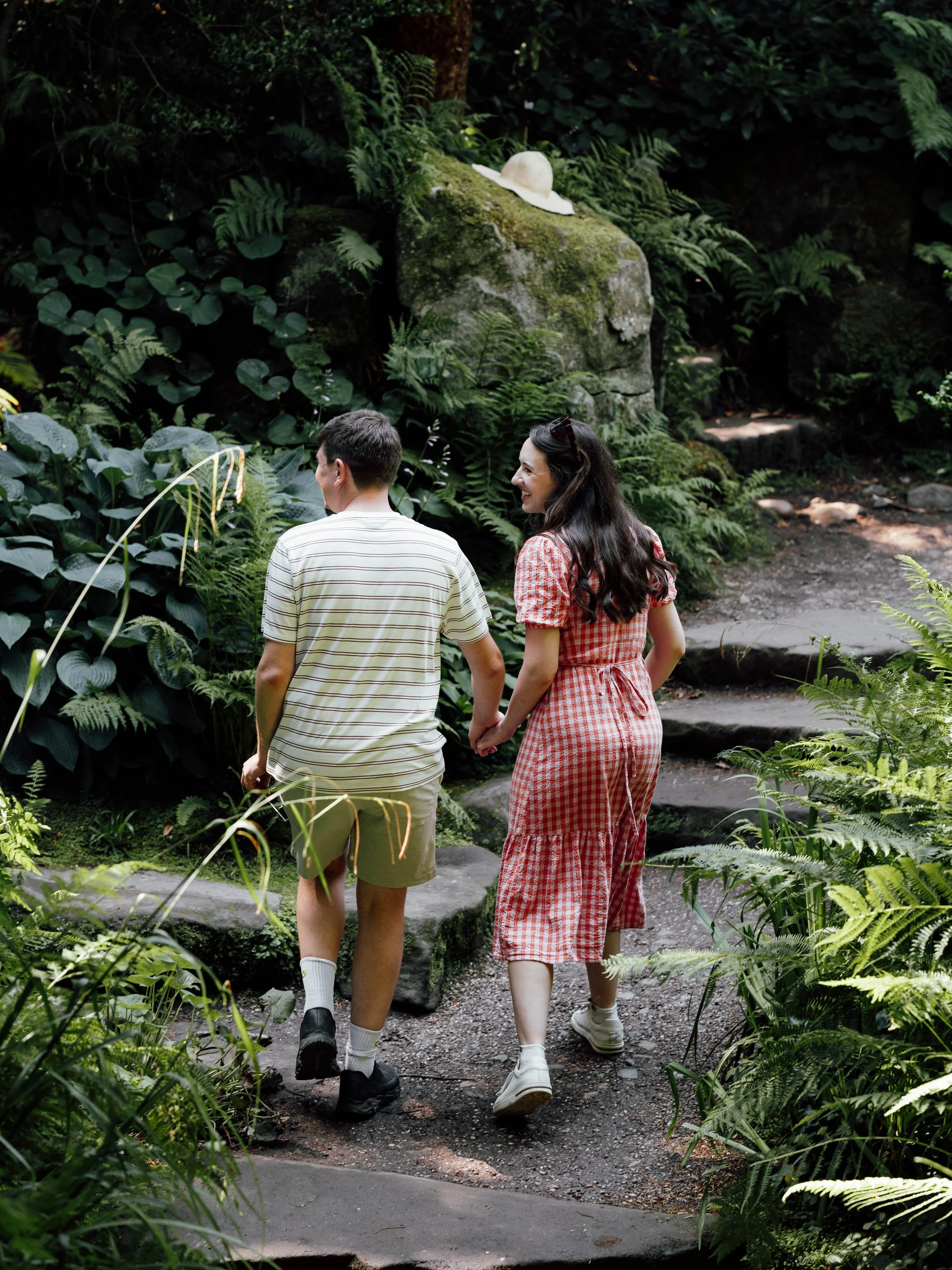 A young couple holding hands and walking on a forest trail surrounded by green plants and ferns.