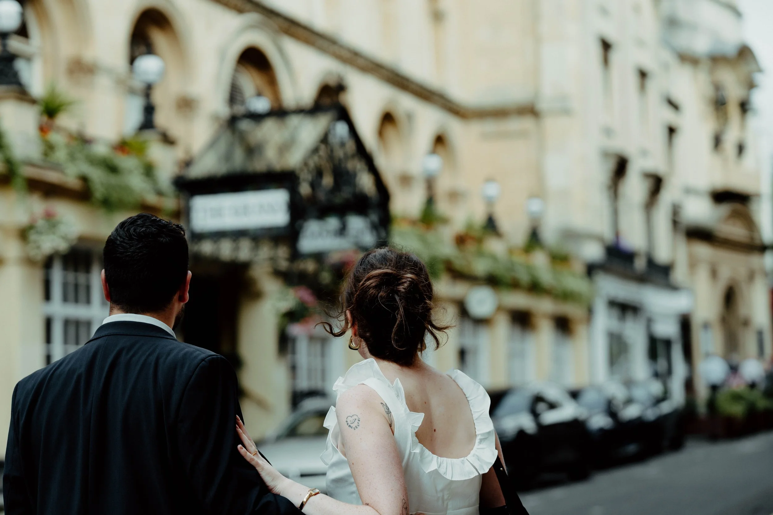A man and woman walk together on a city street, with European-style buildings and parked cars in the background.