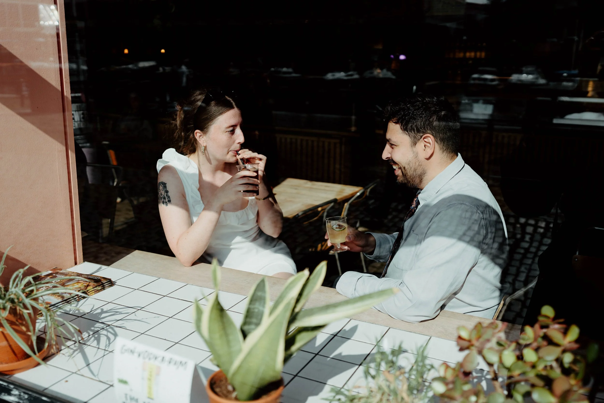 A man and woman are sitting at a cafe window at night, smiling and talking while drinking cocktails. The woman is wearing a white dress and the man a light-colored shirt with a tie. There are potted plants on the counter in the foreground.