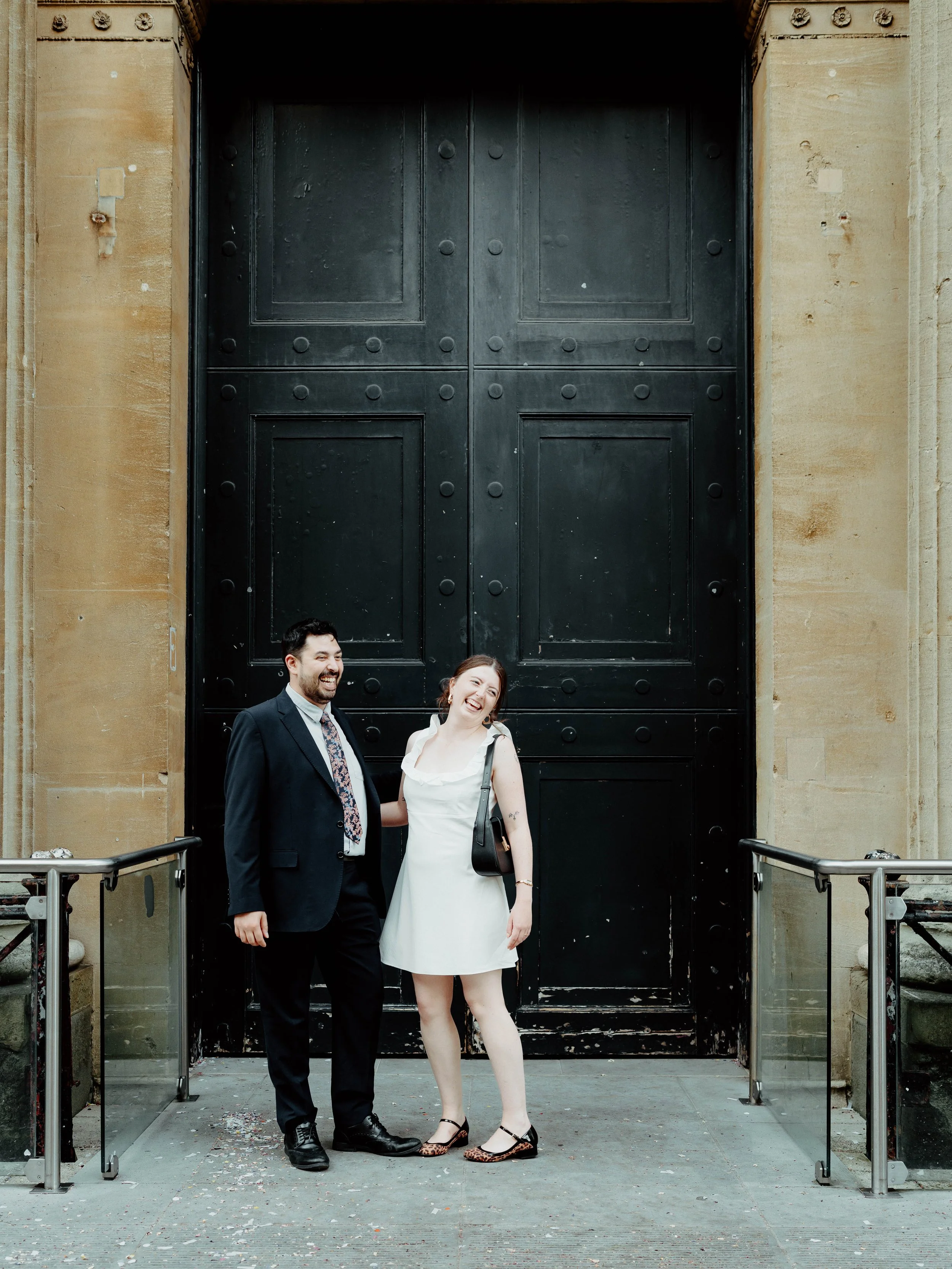 A man and woman standing together in front of a large black door, both smiling and appearing happy.