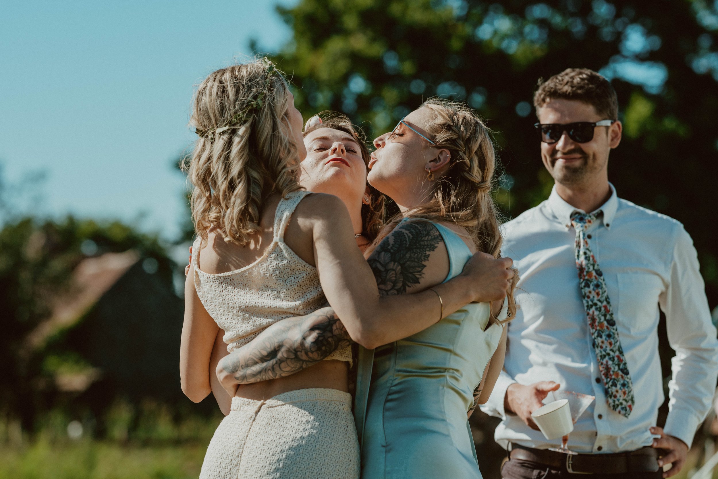 Three women hugging each other outdoors, with a man standing nearby, all smiling and enjoying a sunny day.