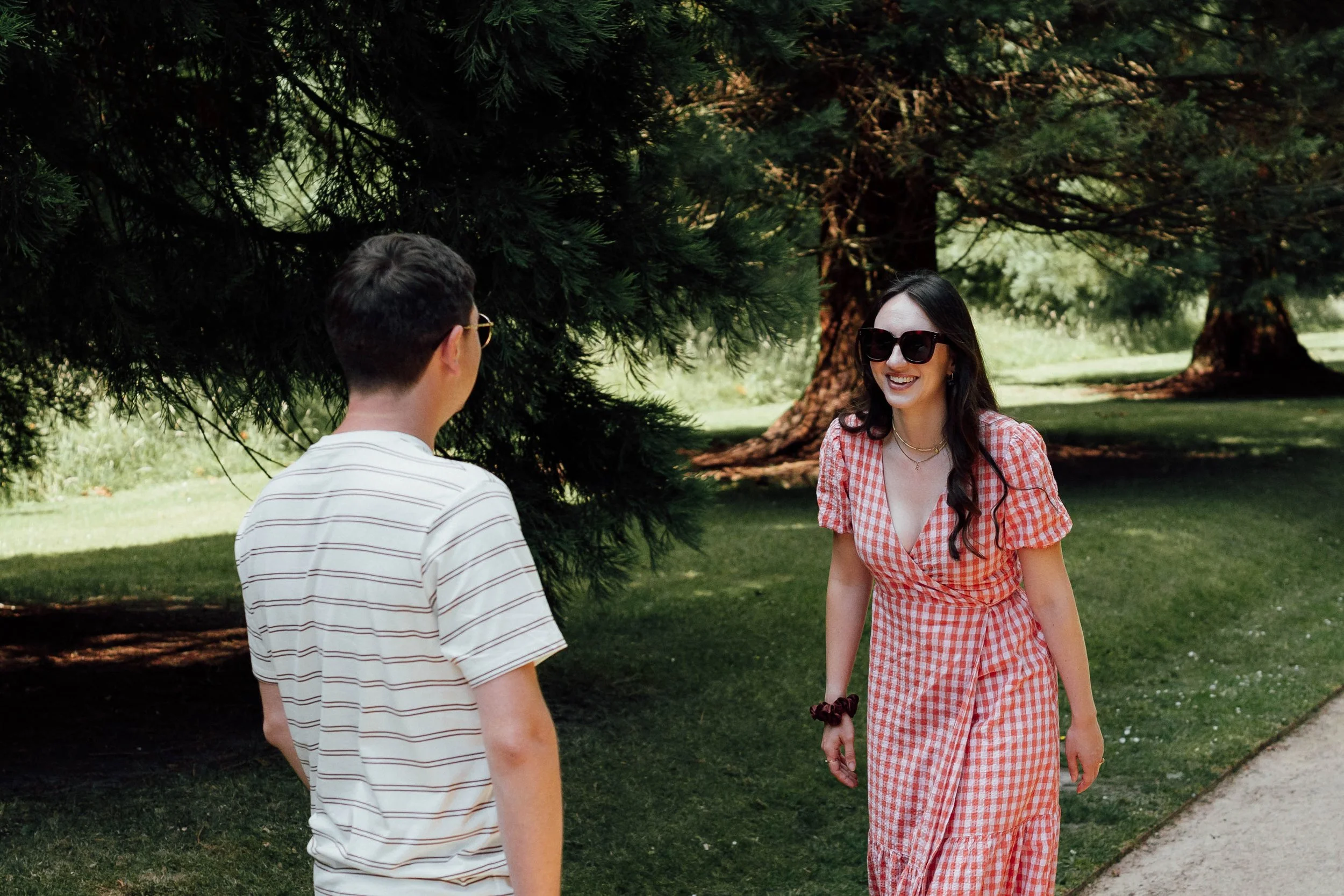 A woman in a red and white checkered dress and black sunglasses is smiling and talking with a man in a striped shirt and glasses outdoors, surrounded by green trees and grass.