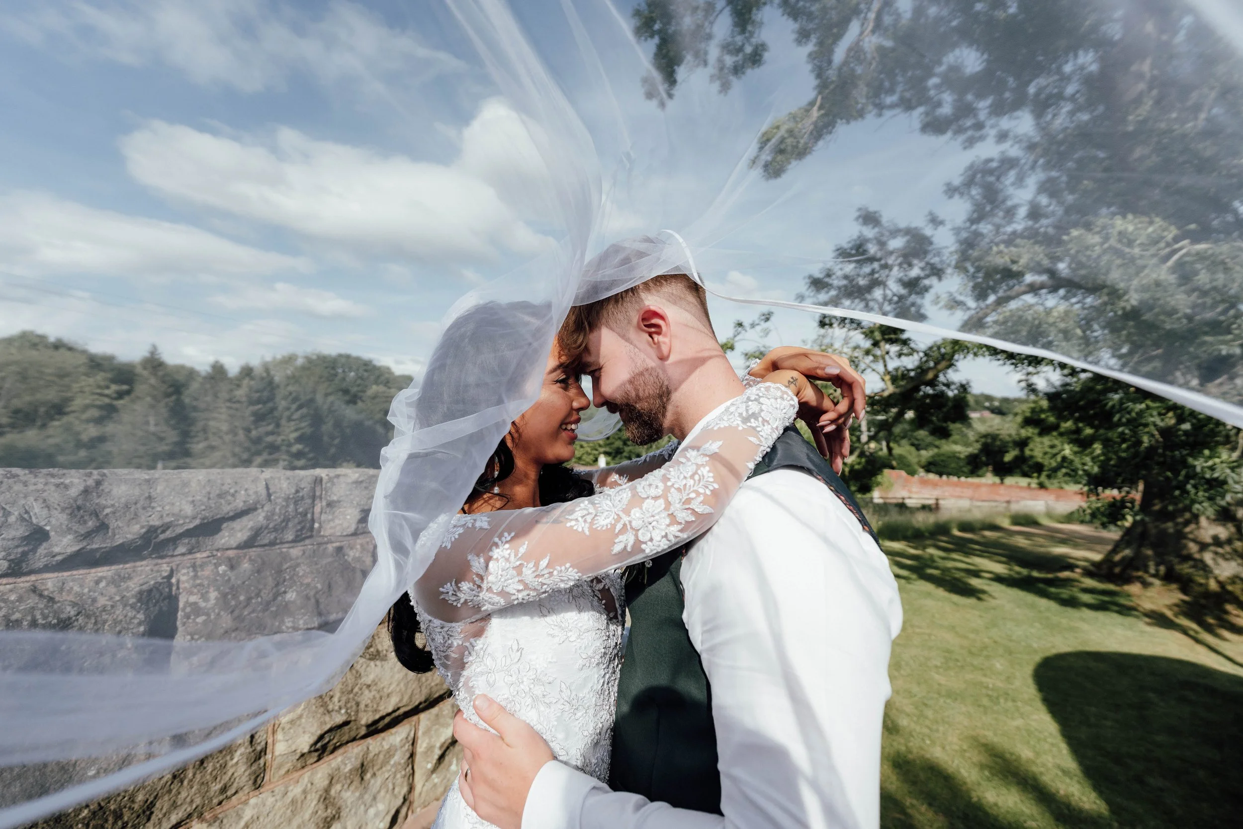 A bride and groom embrace outdoors on a sunny day, with the bride's veil flowing in the wind and trees and a stone wall in the background.