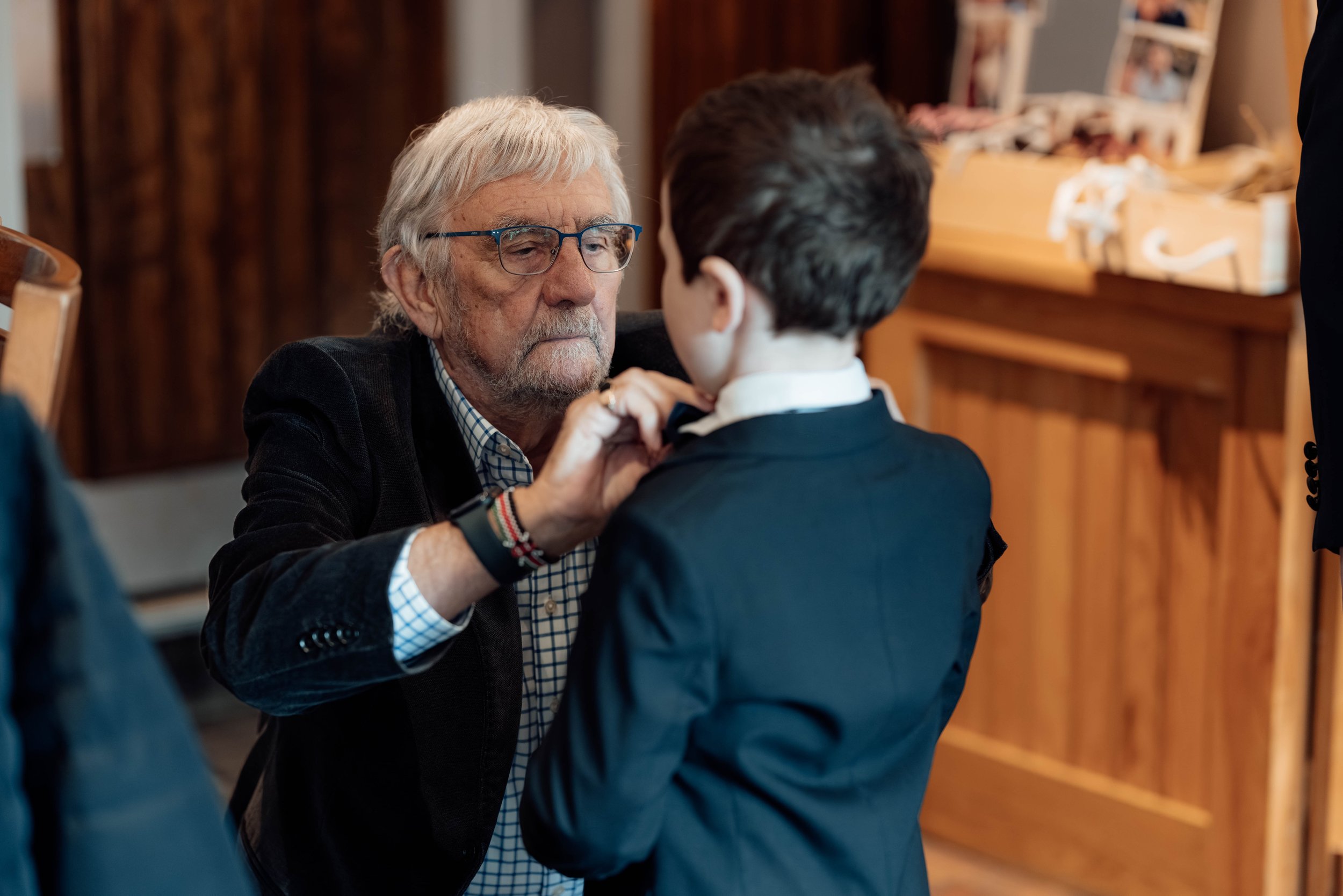 An elderly man with glasses and a beard adjusting a young boy's bow tie in a room with wooden furniture.