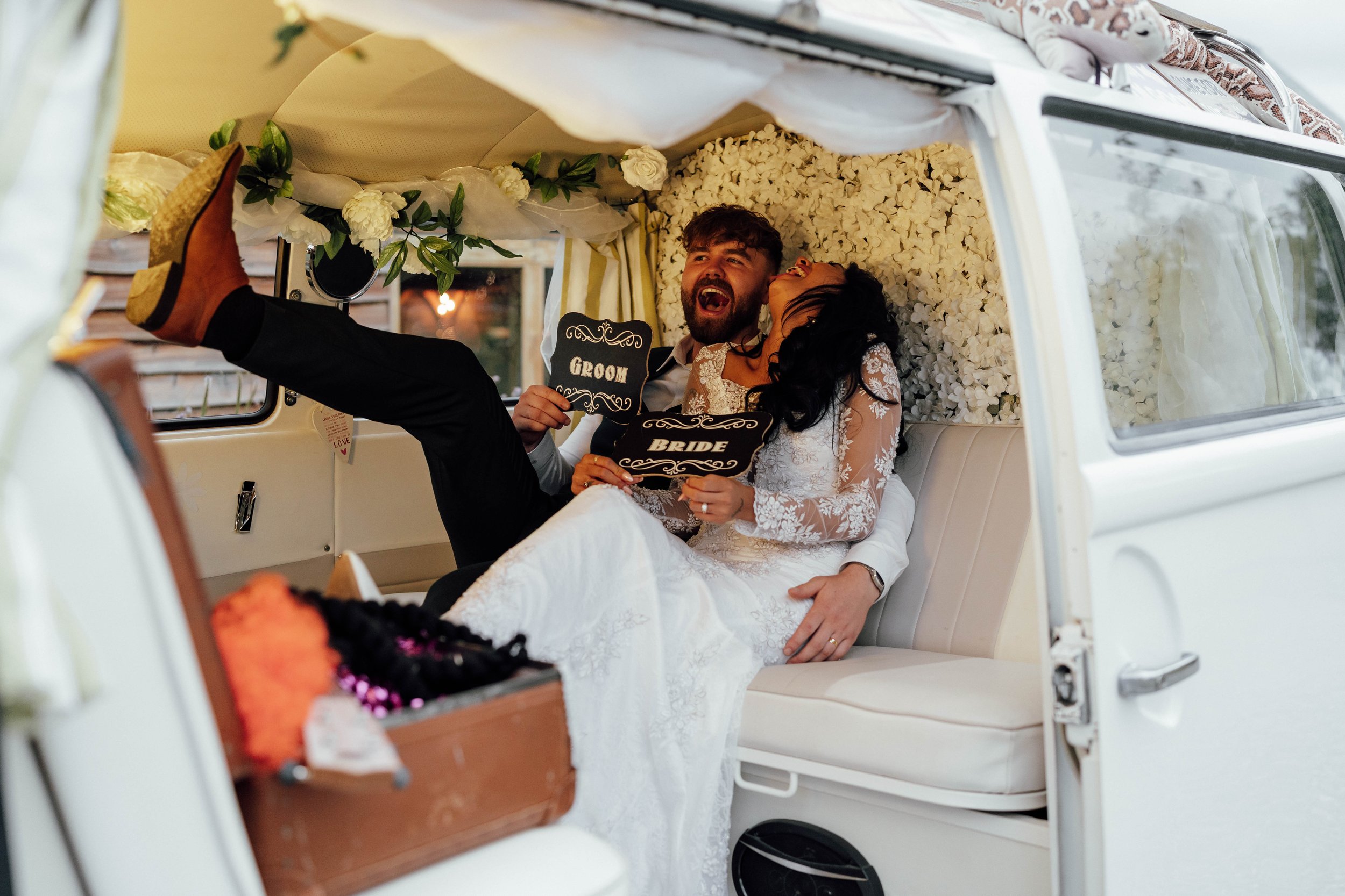 Bride and groom inside a decorated vintage van, smiling and holding signs that say 'Groom' and 'Bride', celebrating their wedding with playful poses.