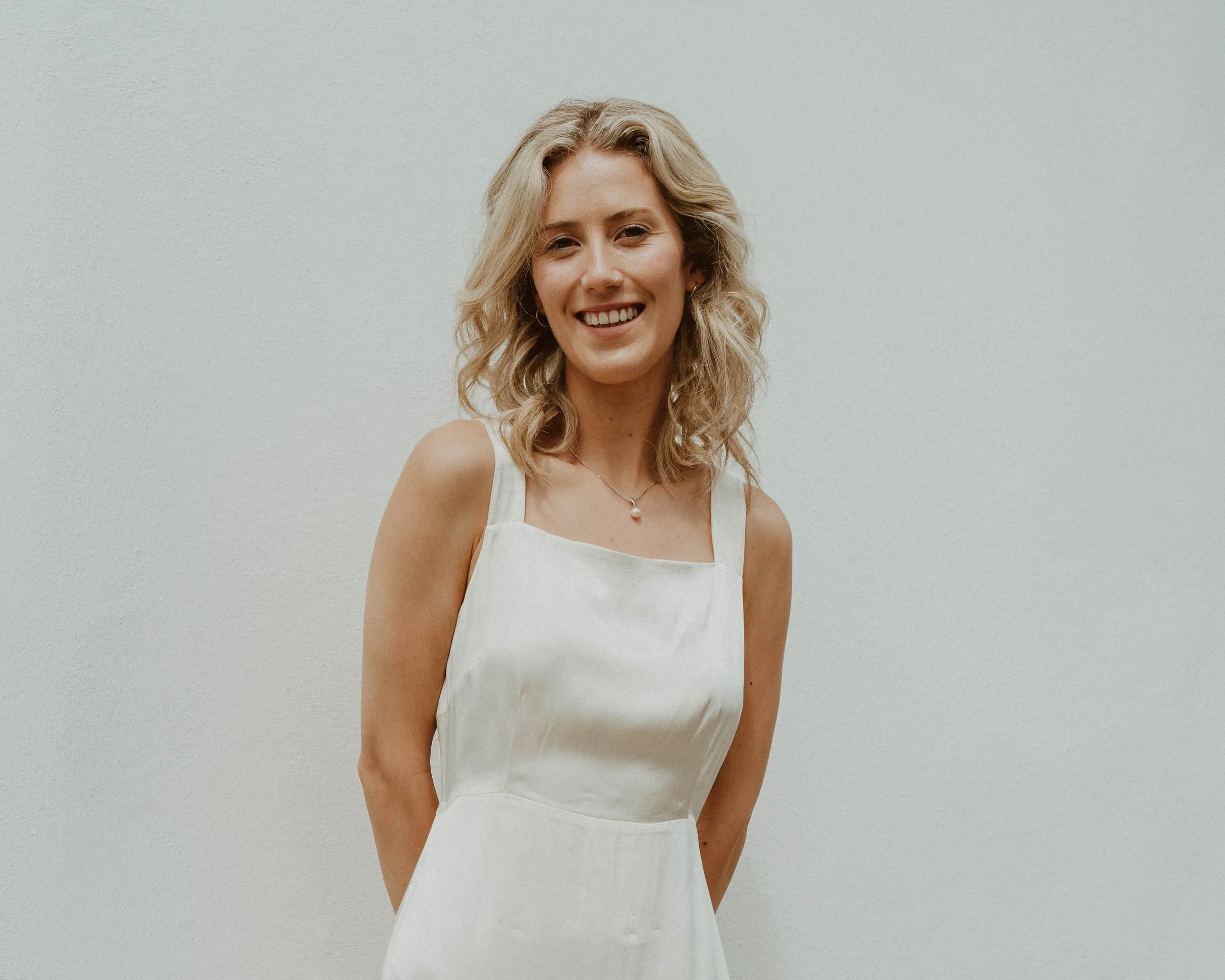 Smiling woman with blonde curly hair in a white sleeveless dress standing against a plain light-colored wall.