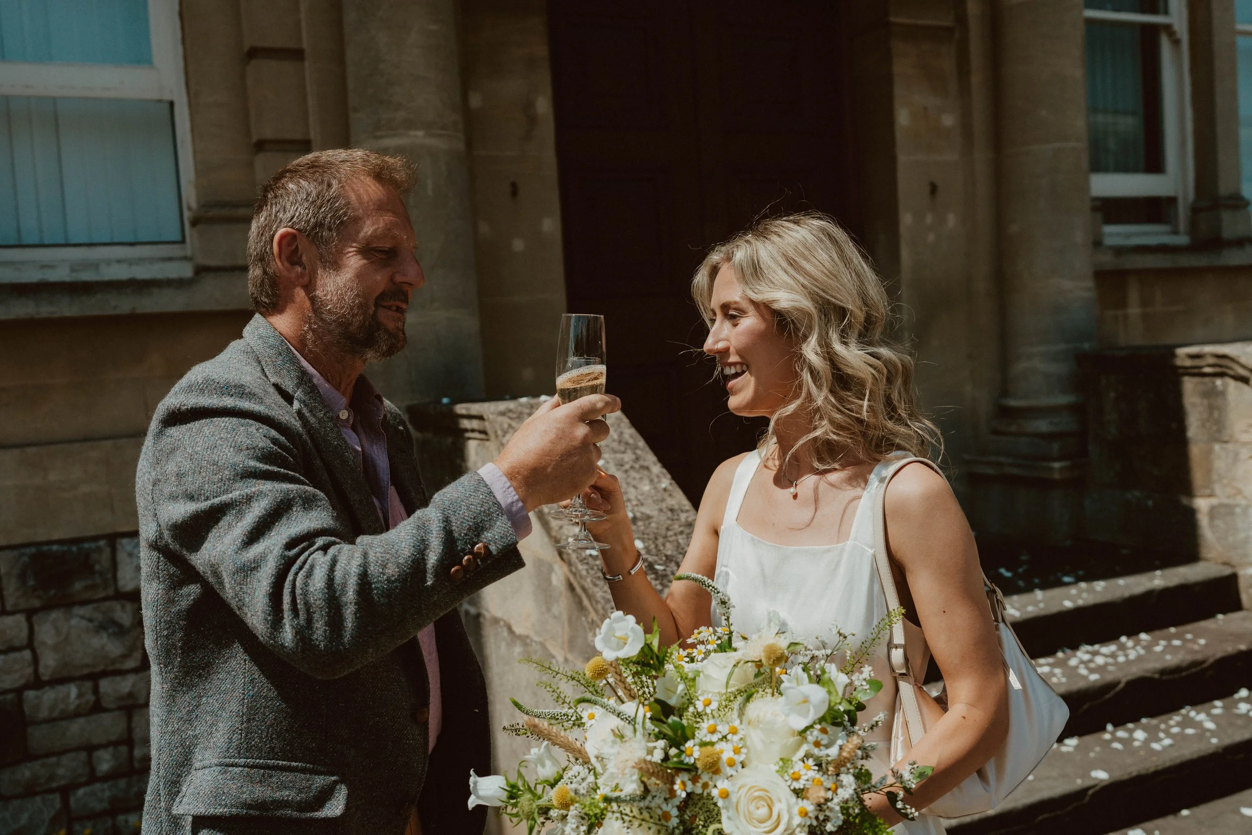 A man in a grey blazer raises a glass of champagne to toast a woman holding a bouquet of flowers, both smiling.