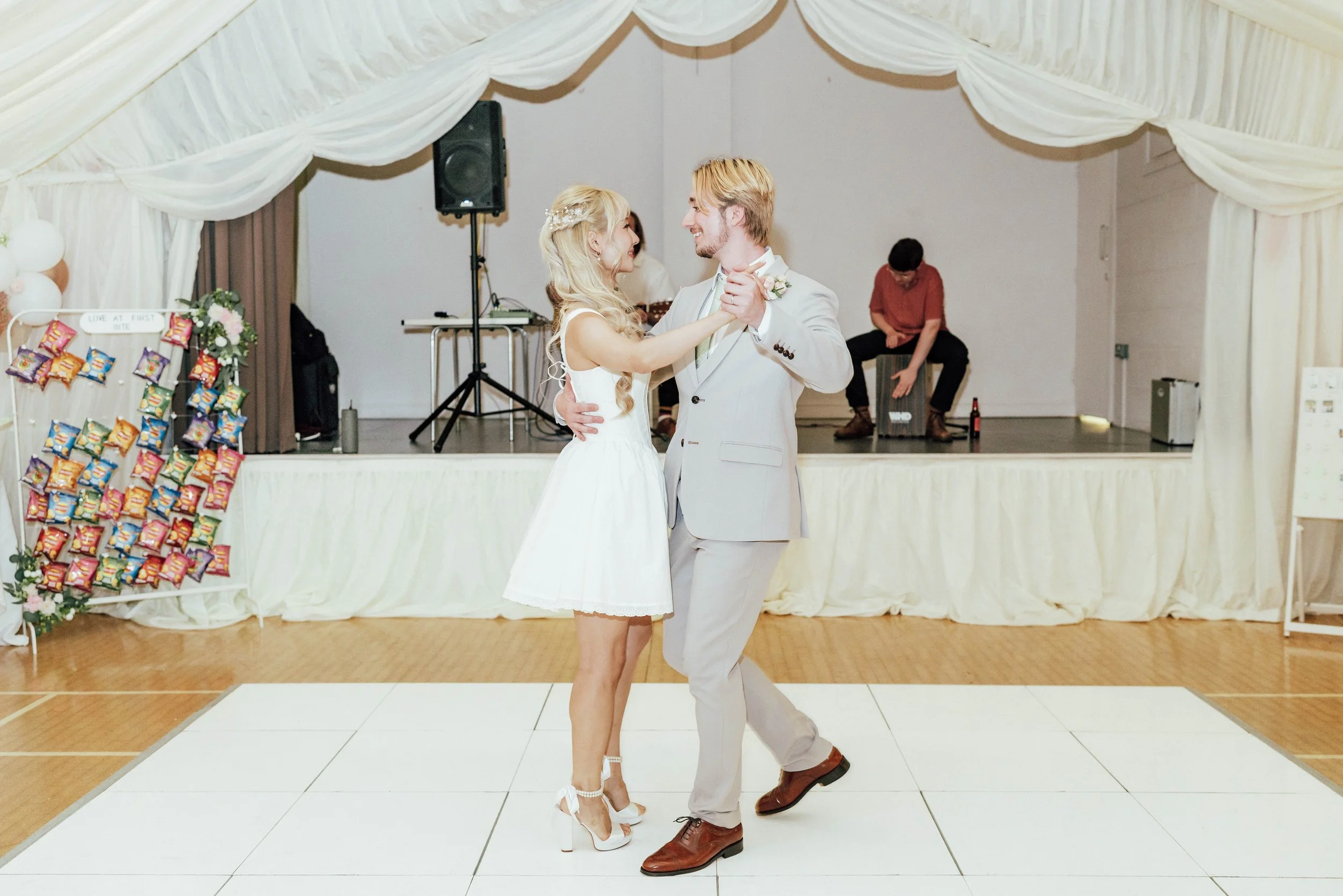 A bride and groom are dancing on a white dance floor at their wedding reception. The bride is in a white dress with white high-heeled shoes, and the groom is in a light gray suit with brown shoes. Behind them, a musician is sitting on a cajón, and th