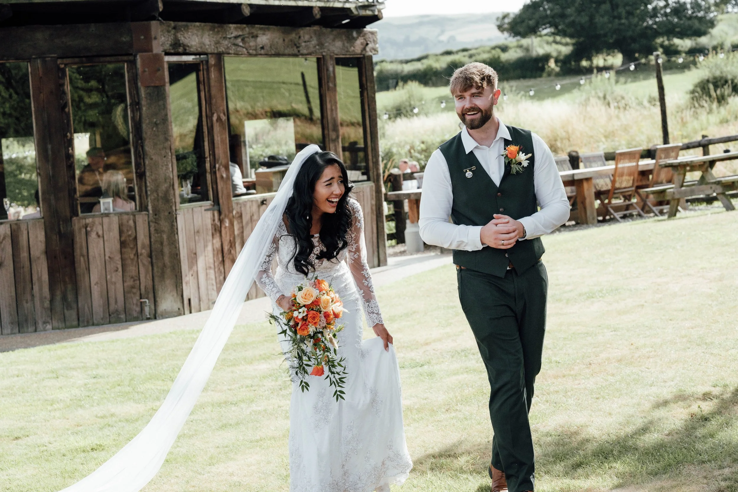 A bride in a white lace wedding dress holding a bouquet of orange, peach, and white flowers, laughing, walking outdoors with a groom in a white shirt and green vest, holding hands, during a wedding celebration on a grassy area with a wooden building 