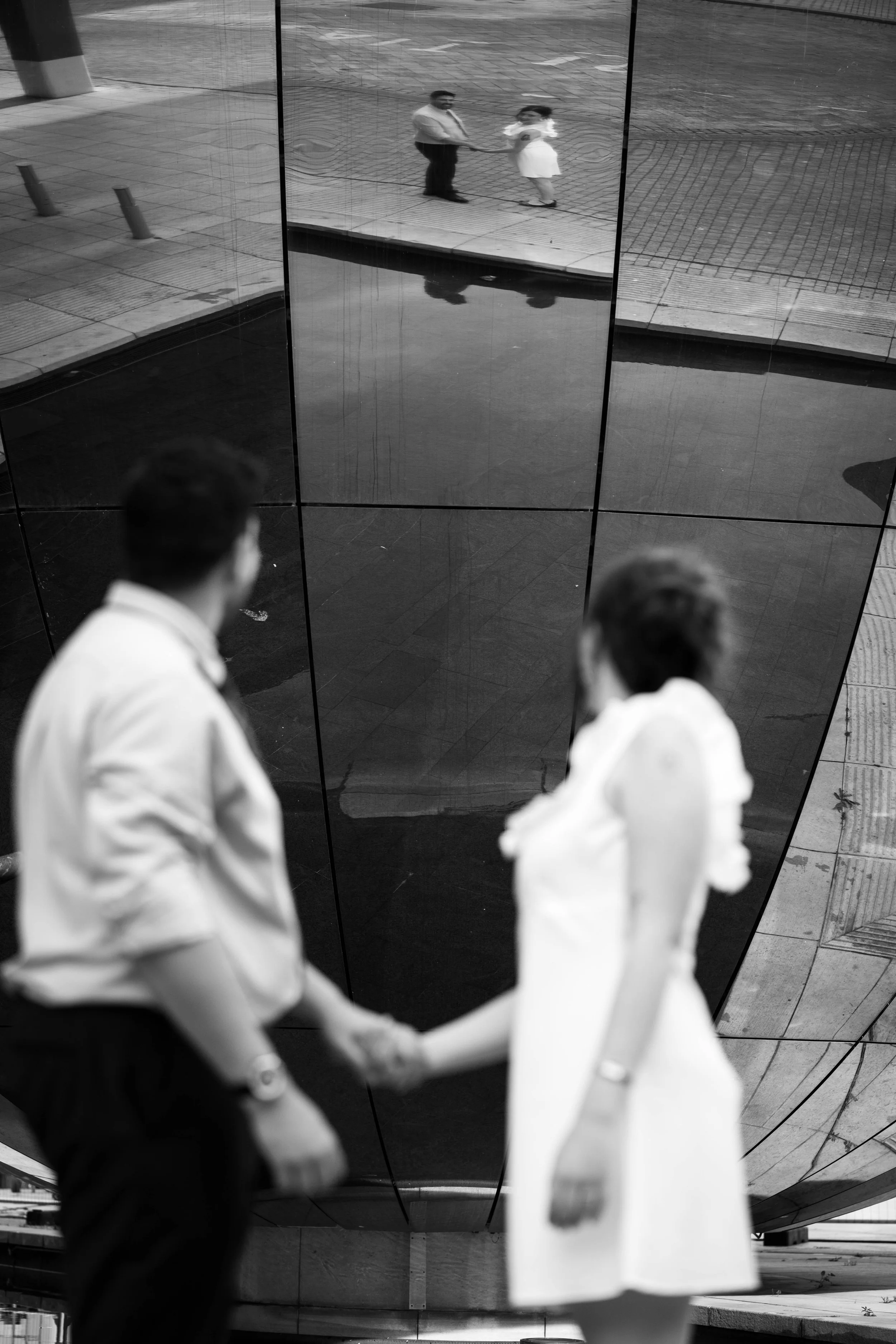 A black and white photo of a man and woman holding hands, standing in front of a reflective metallic surface, with their reflection visible in it.